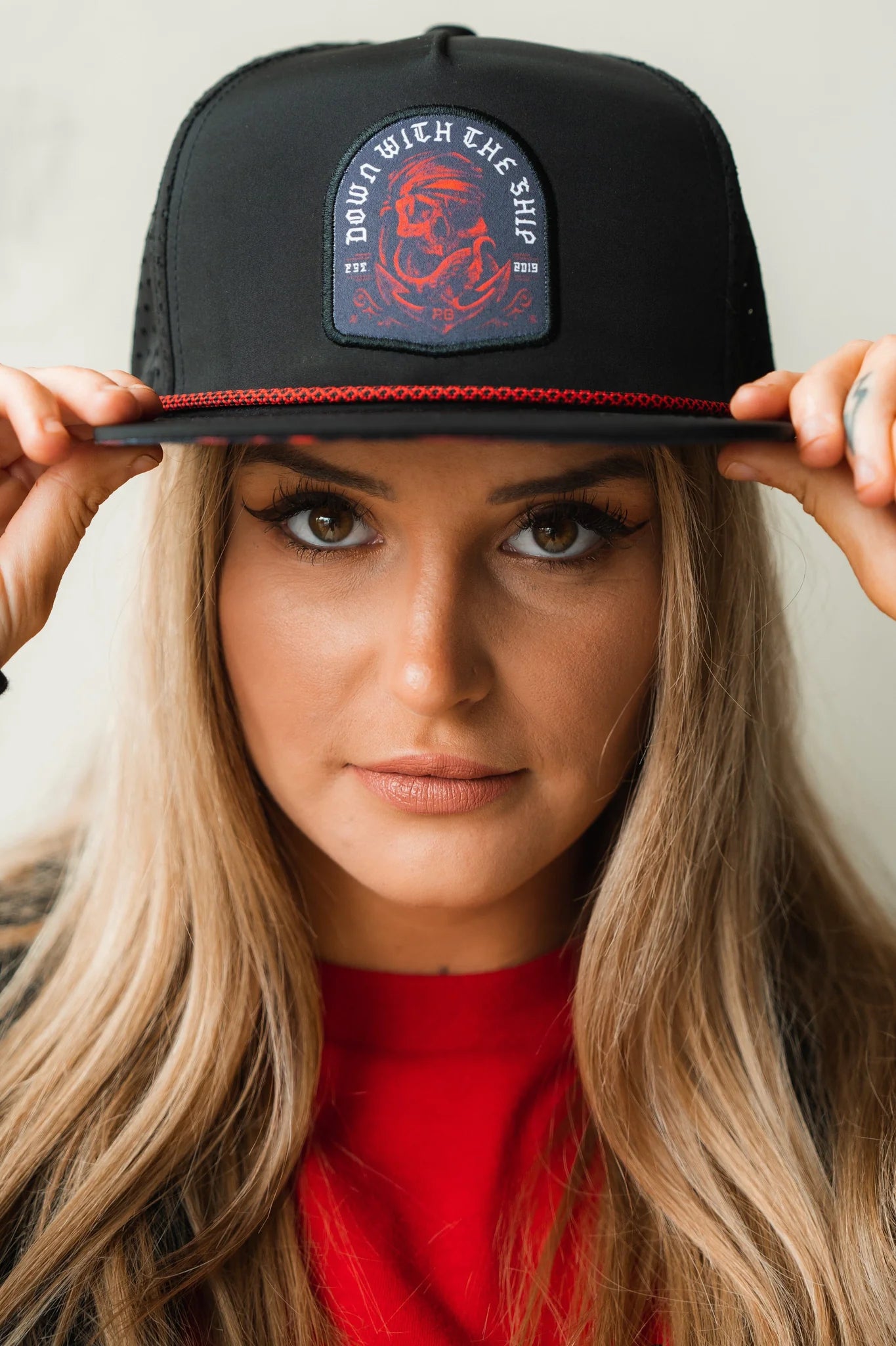 A person with long blonde hair wears the handcrafted Ship Wreck Snapback - Red by Relentless Betrayal, featuring a red and white DOWN WITH THE SHIP embroidered patch. The high-quality hat sits snugly on their head as they hold its brim, making eye contact with the camera in a red top.