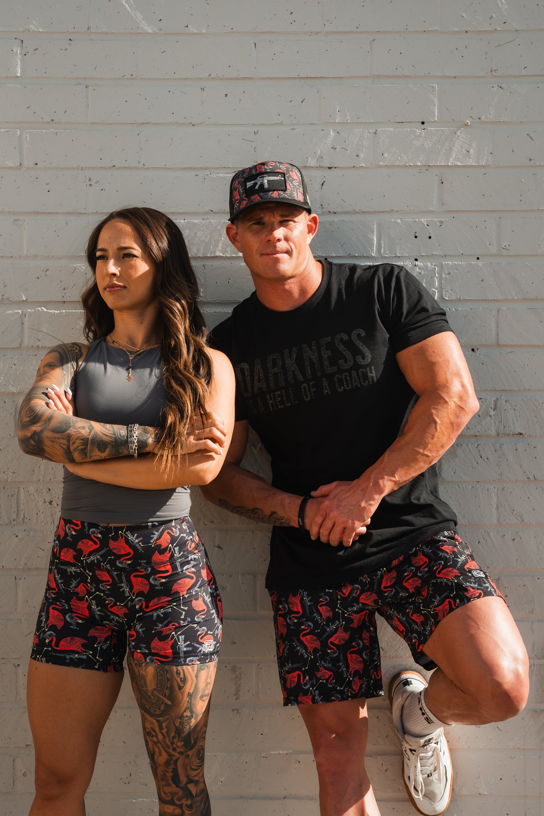Two people stand against a white brick wall. On the left, one wears a gray top and patterned shorts, showing off their tattoos with style in breathable fabric. The person on the right sports a black cap, dark shirt, and Tango Charlie Apparels Womens High-Waisted Seamless Front Compression Training Shorts | Tactimingo.