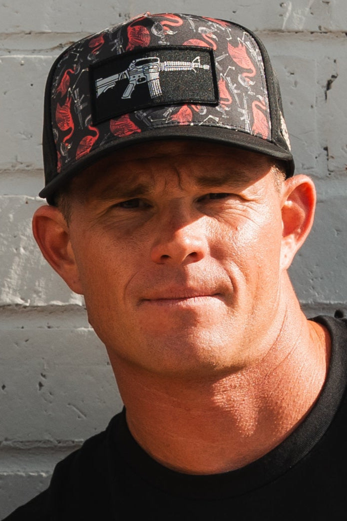 A man stands against a white brick wall, wearing a black T-shirt and a Tango Charlie Apparel Tactimingo Hydro Snapback Hat adorned with a rifle graphic patch.