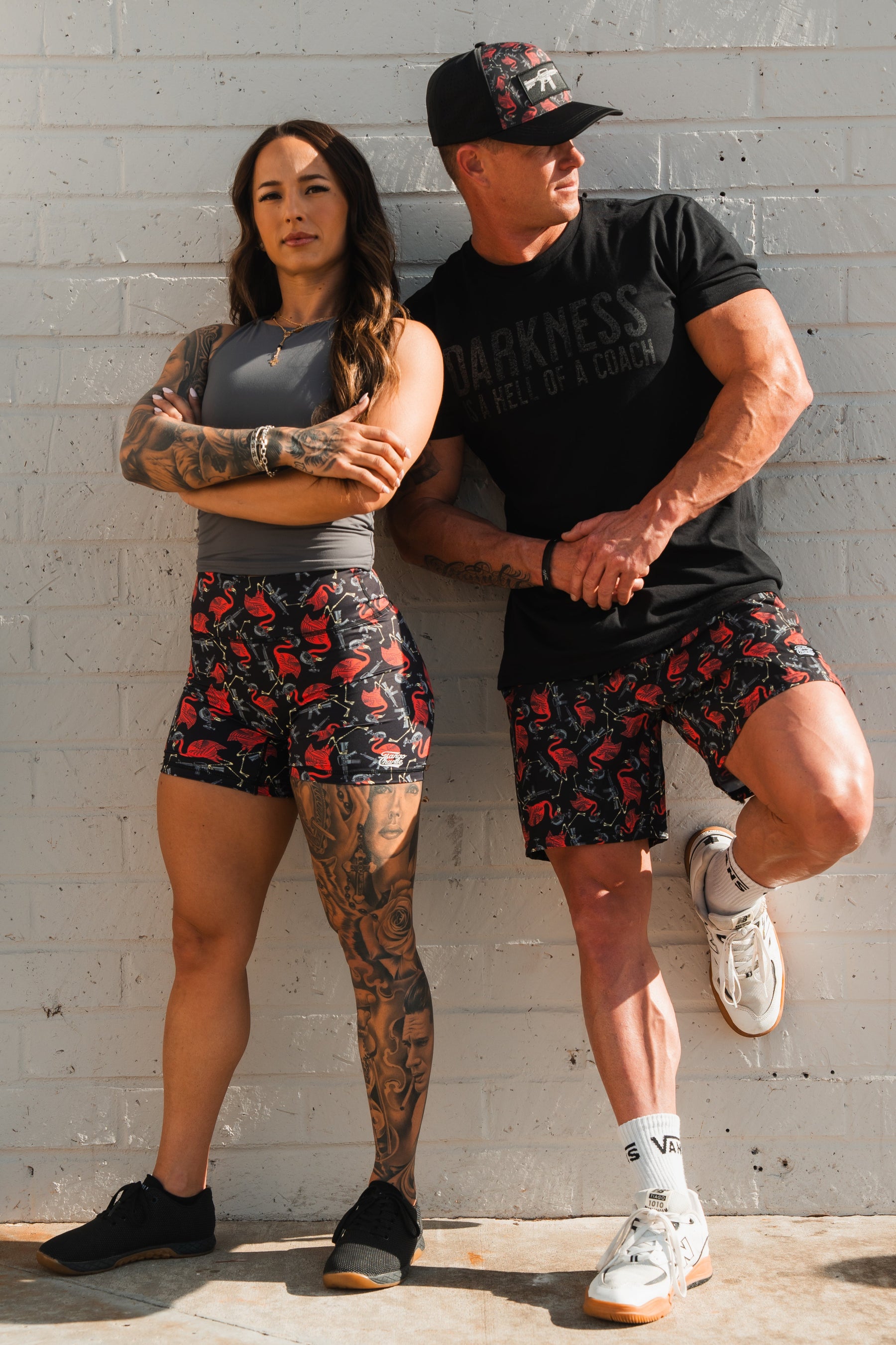 Two athletic individuals stand against a white brick wall. The woman wears a gray top and Womens High-Waisted Seamless Front Compression Training Shorts by Tango Charlie Apparel, with tattoos on her arm and leg. The man sports a black shirt, matching shorts, and cap with a similar pattern in relaxed poses.