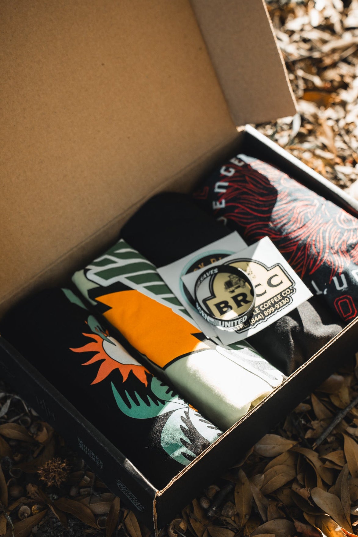 An open Born United Private Label Born United Black Box with folded mystery tees and a BRCC sticker inside, placed on dried leaves outdoors.