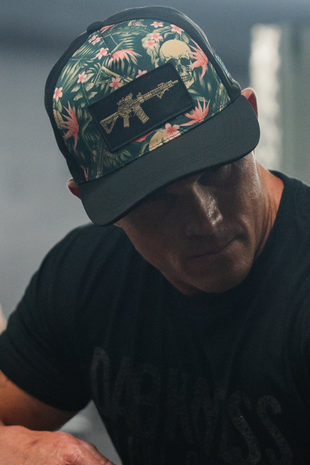 The person wears a sleek black t-shirt and a Tango Charlie Apparel Aloha AR Hydro Snapback Hat with a tropical design, featuring a rifle patch. The blurred background highlights their cool style as shadows partly obscure their face.