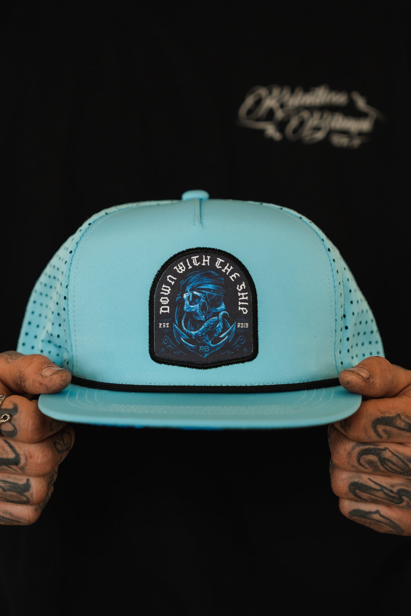 A person with tattooed hands holds a Ship Wreck Snapback - Blue by Relentless Betrayal, featuring a skull patch and DOWN WITH THE SHIP text. They wear a black shirt with white lettering in the background.