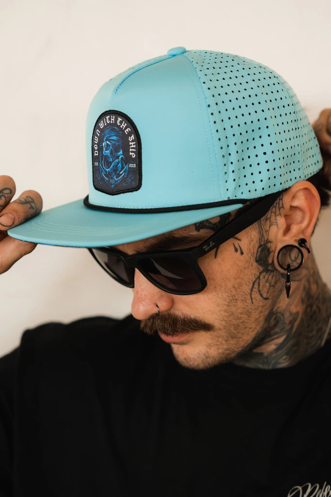 With tattoos and ear piercings, a person sports a mustache, sunglasses, and a Relentless Betrayal Ship Wreck Snapback in blue with a nautical patch. They adjust the breathable cap while wearing a black shirt against a plain background.