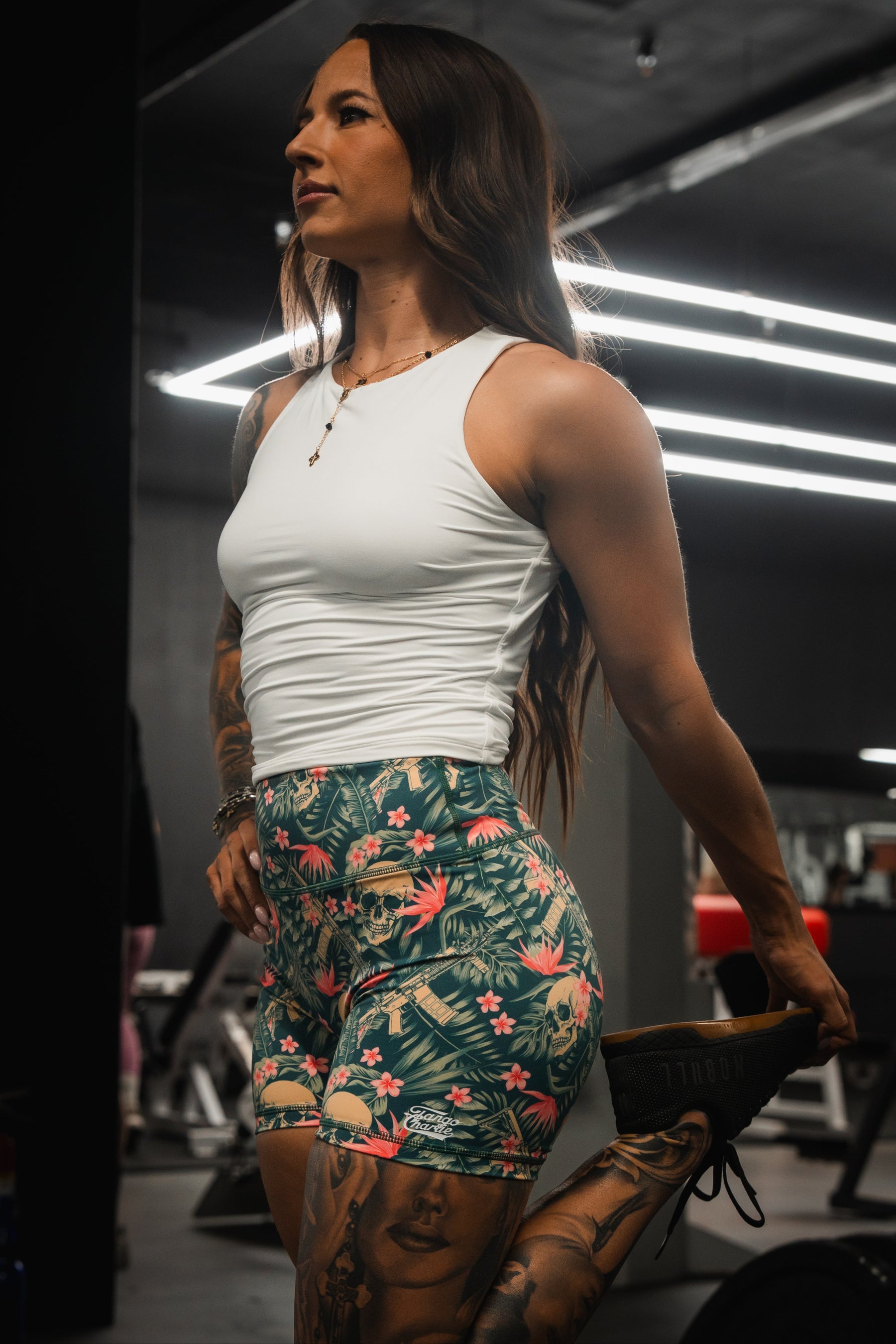 In a gym, a woman in Tango Charlie Apparels Womens High-Waisted Seamless Front Compression Training Shorts by Aloha AR stretches her leg behind her. Her long hair cascades, revealing limb tattoos amid the gears empowering design under bright lights surrounded by equipment.