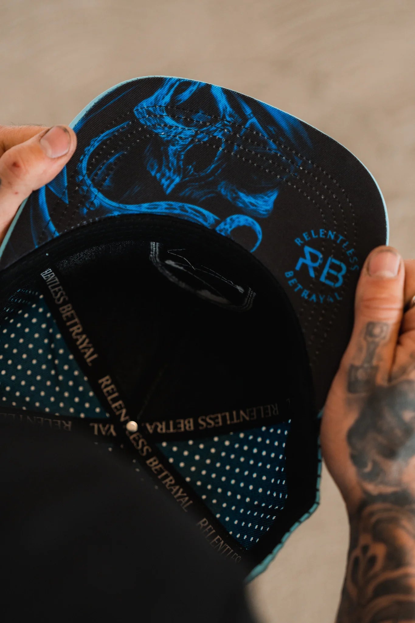 A person with tattooed hands holds a breathable snapback, revealing the underside of the brim. The Ship Wreck Snapback - Blue by Relentless Betrayal features RB initials and a stylish polka dot pattern inside.