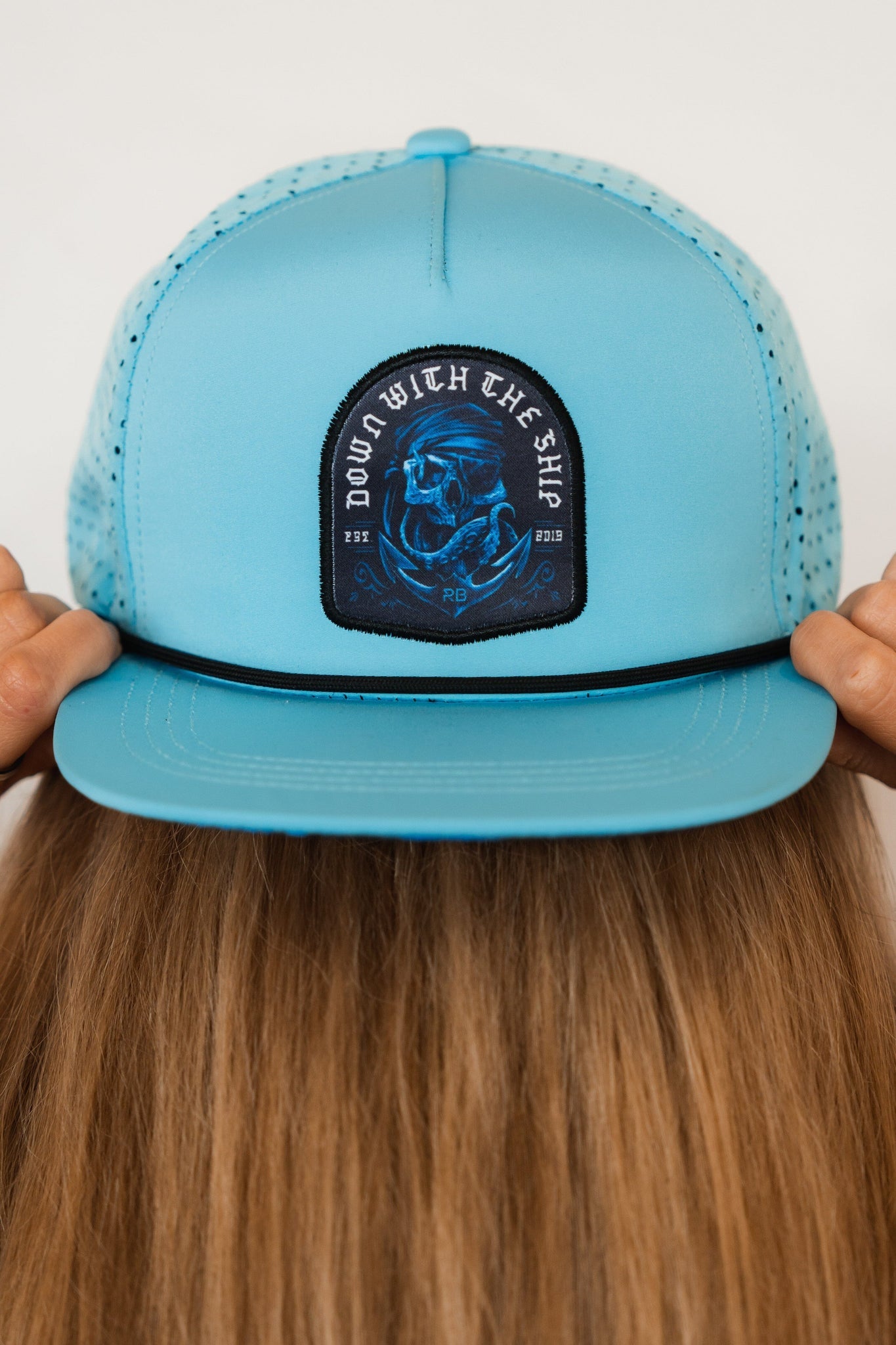 A person with long brown hair is wearing the light blue Ship Wreck Snapback by Relentless Betrayal. This breathable cap features a skull and bones patch with the text DOWN WITH THE SHIP, evoking high-seas adventures as their hands adjust it from behind.