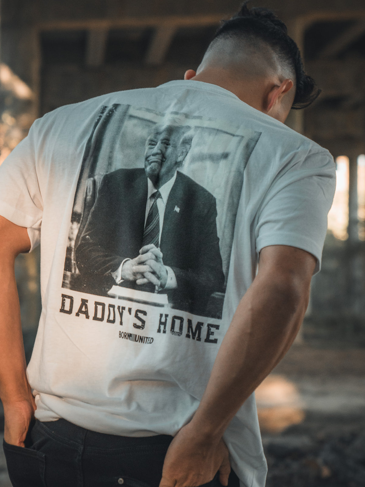 Outdoors and partially turned away, a person wears the Daddys Home - #47 - White tee by Born United Private Label, featuring a suited man with clasped hands and hinting at Trump’s second win.