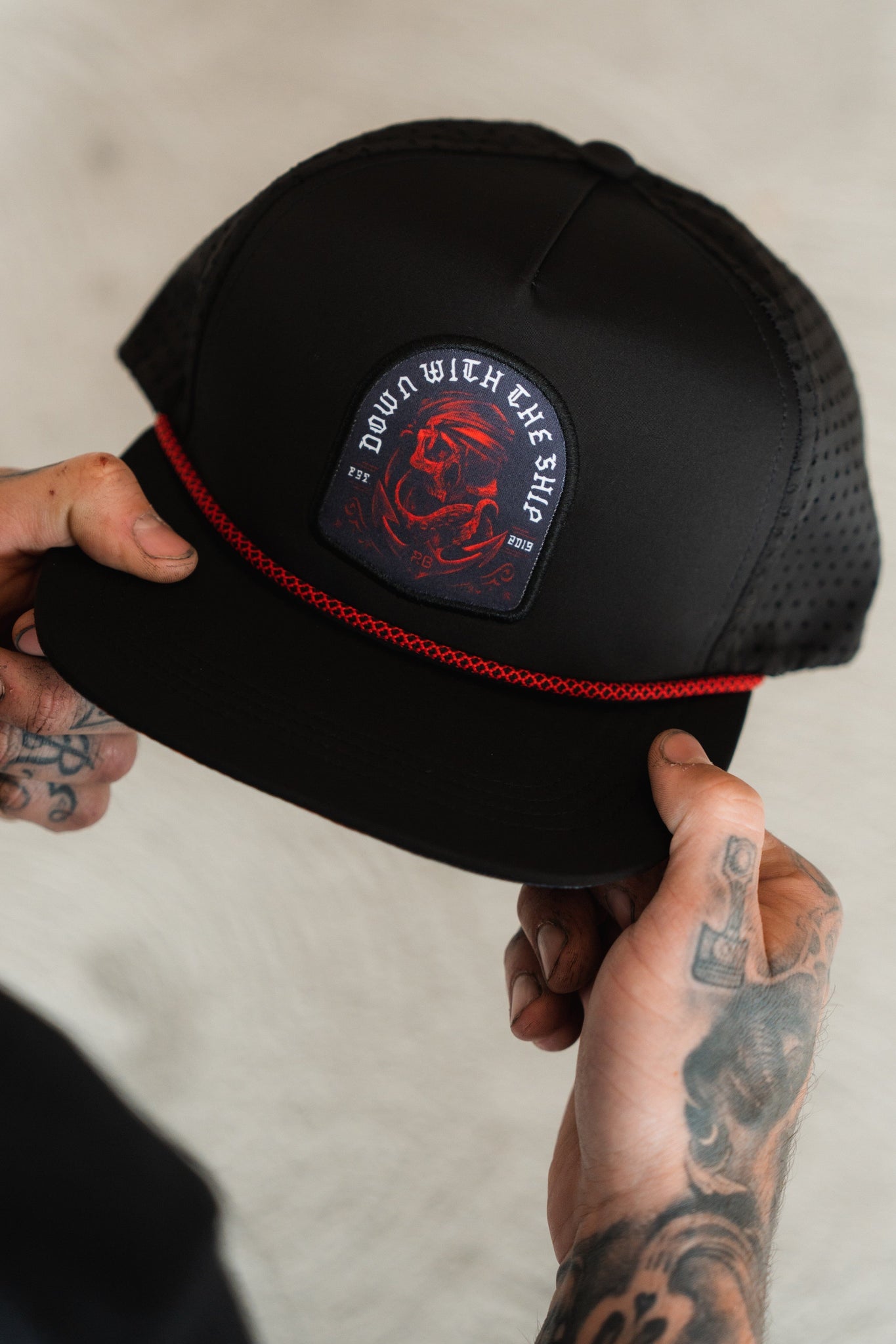 A person with tattooed hands holds the Ship Wreck Snapback - Red by Relentless Betrayal, featuring a red dragon and DOWN WITH THE SHIP text, textured mesh sides, and a red stripe above the brim.