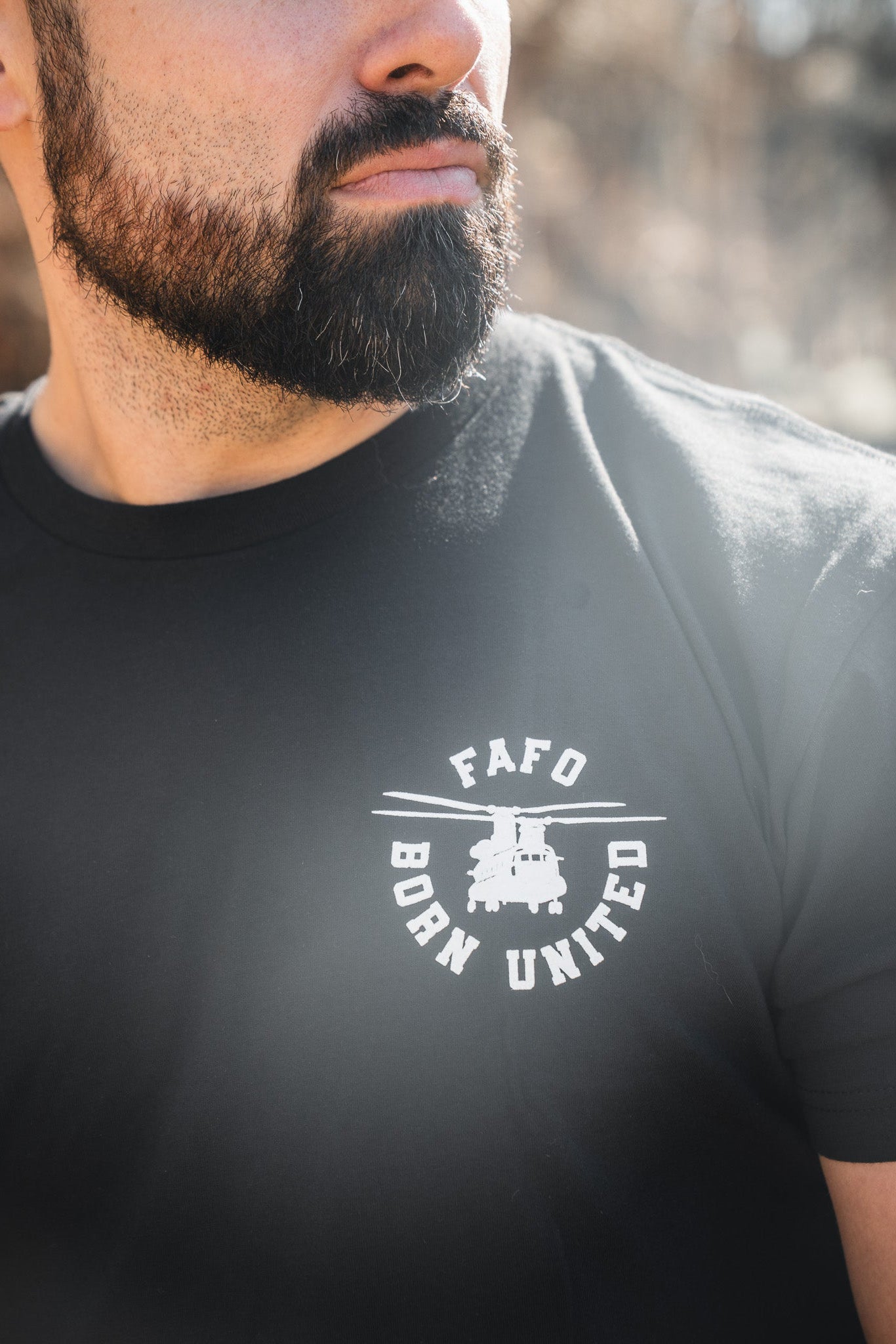 A bearded man wears the FAFO – Maduro black tee by Born United Private Label, featuring a white logo and small vehicle graphic. This limited design is seen outdoors with sunlight highlighting its bold style.