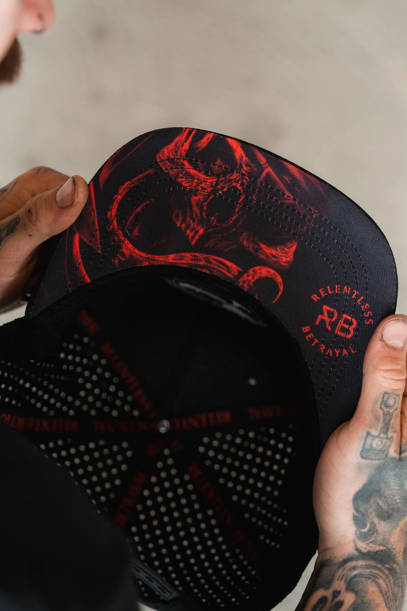 Tattooed hands display the Ship Wreck Snapback - Red from Relentless Betrayal, featuring a black cap with a red demonic illustration under the brim and adorned with RELENTLESS BETRAYAL and RB text for those who love unique style.