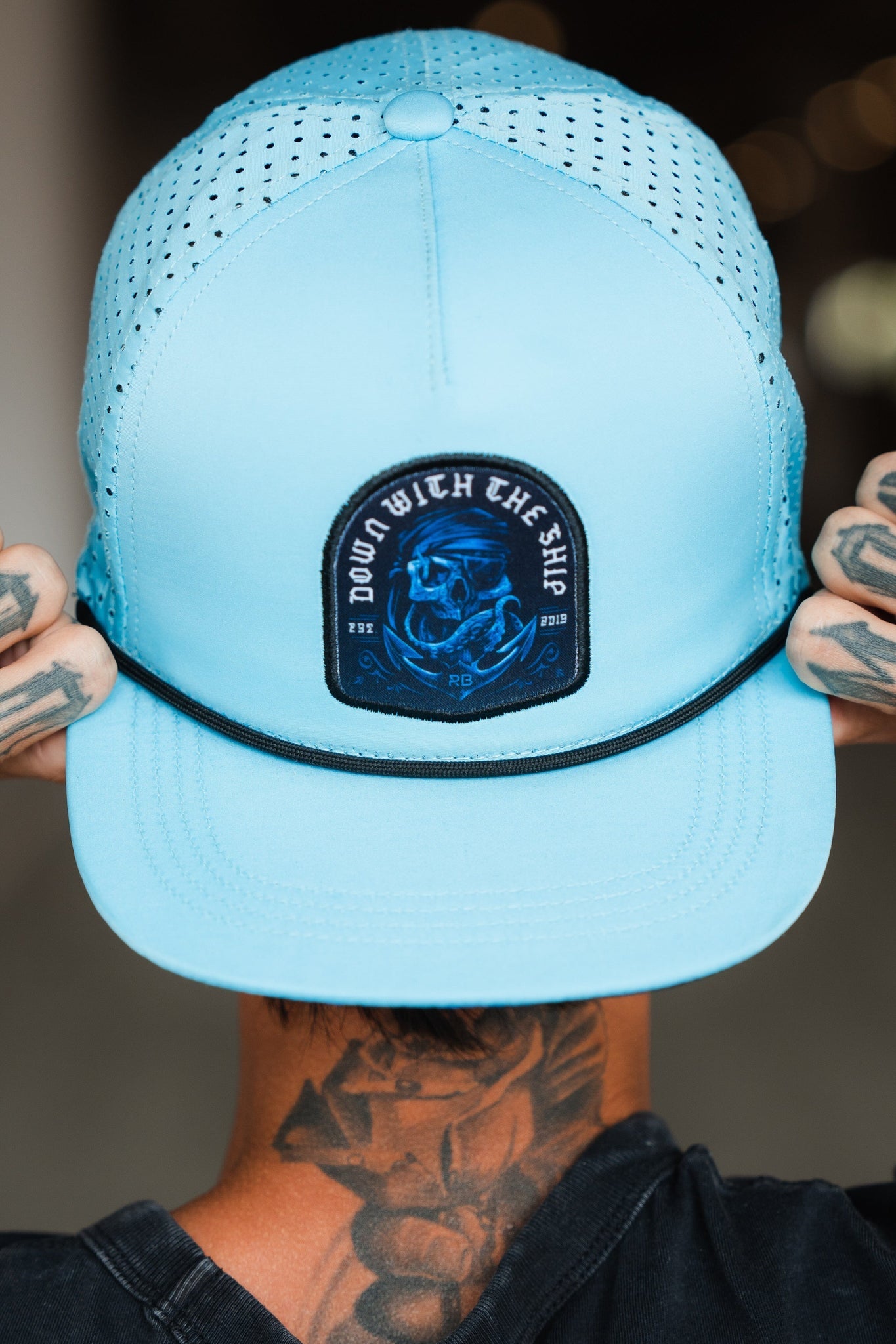 A tattooed person adjusts the brim of a Ship Wreck Snapback - Blue by Relentless Betrayal, showcasing its light blue color, black DOWN WITH THE SHIP patch, and breathable perforations.