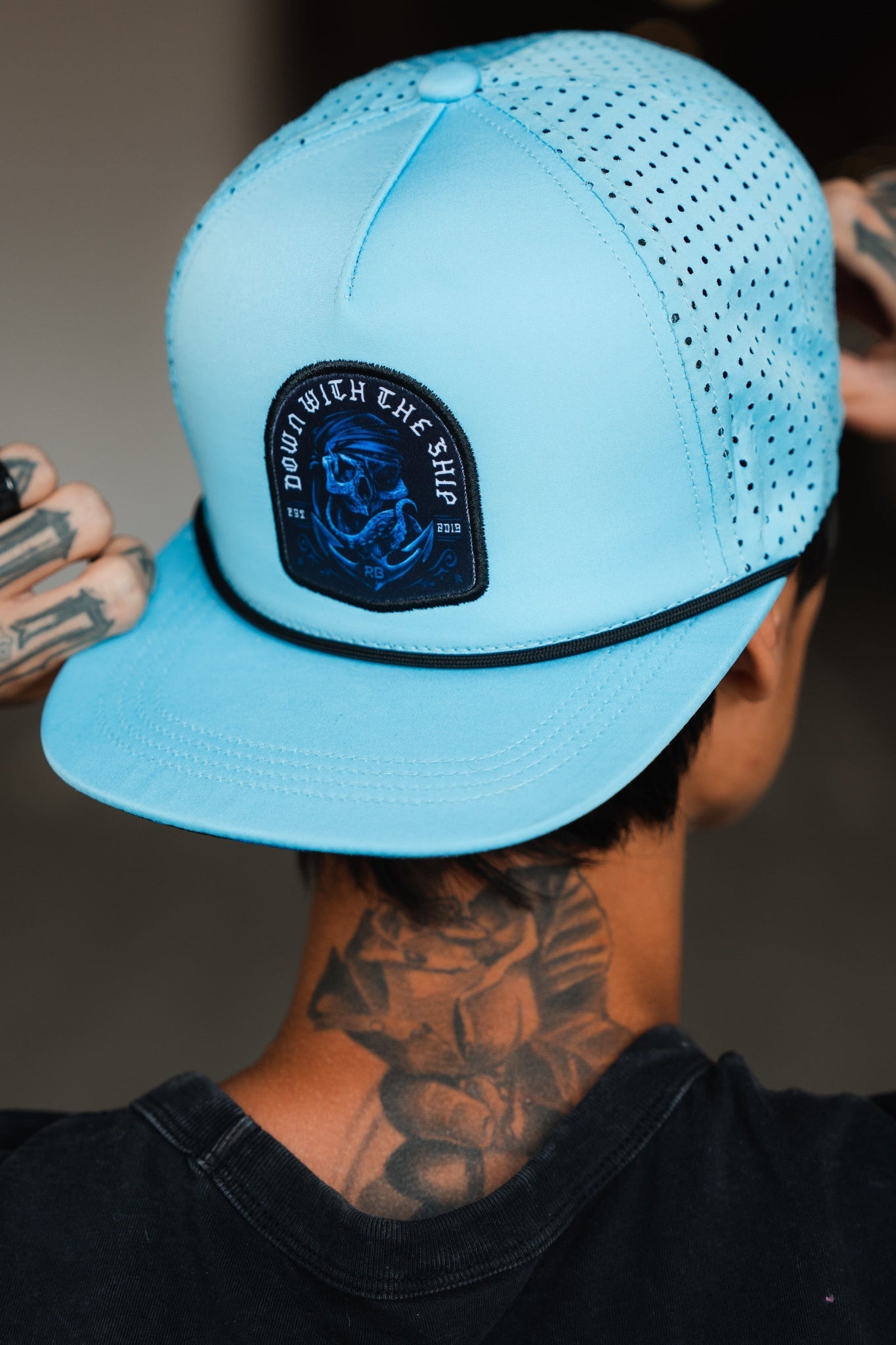 Wearing the Ship Wreck Snapback in blue by Relentless Betrayal, a person adjusts the breathable cap with its skull and text black patch. A bold neck tattoo adds intrigue against a blurred backdrop.