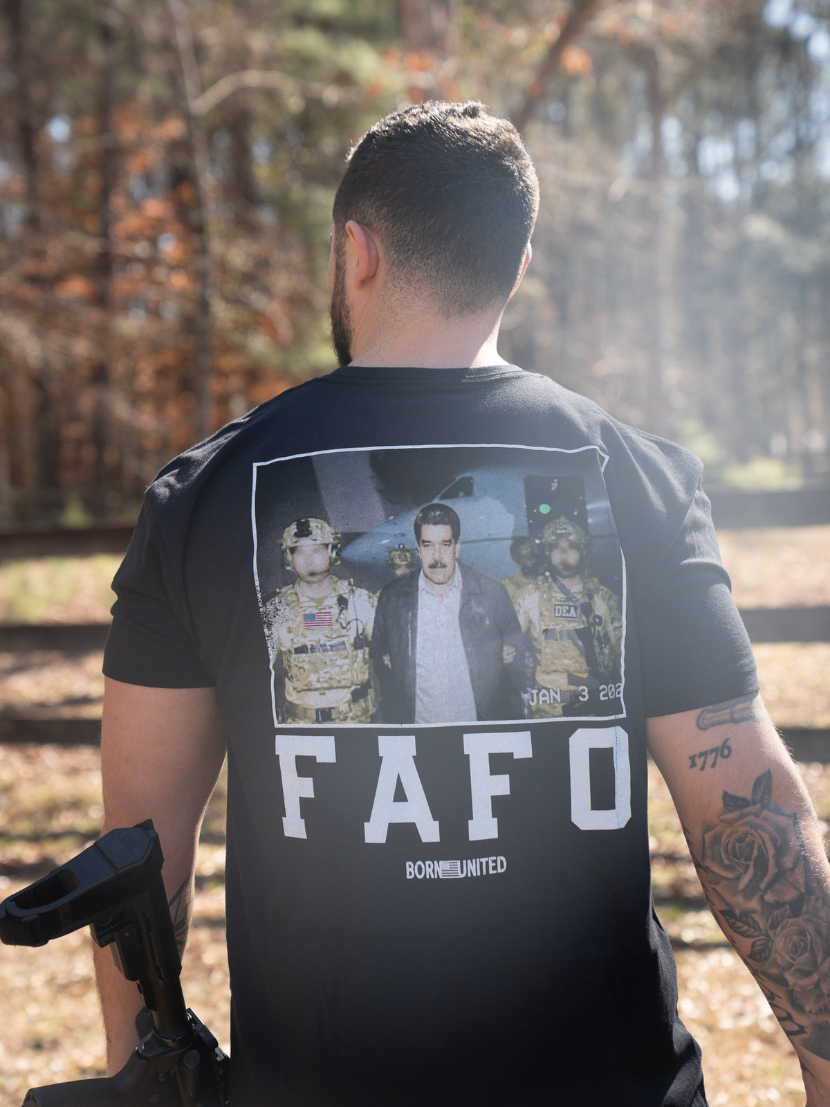 A man stands outdoors with his back to the camera, wearing the Born United Private Label FAFO - Maduro T-shirt, which features a photo of an arrest scene and bold FAFO and BORN UNITED text. Sunlight filters through the trees behind him.