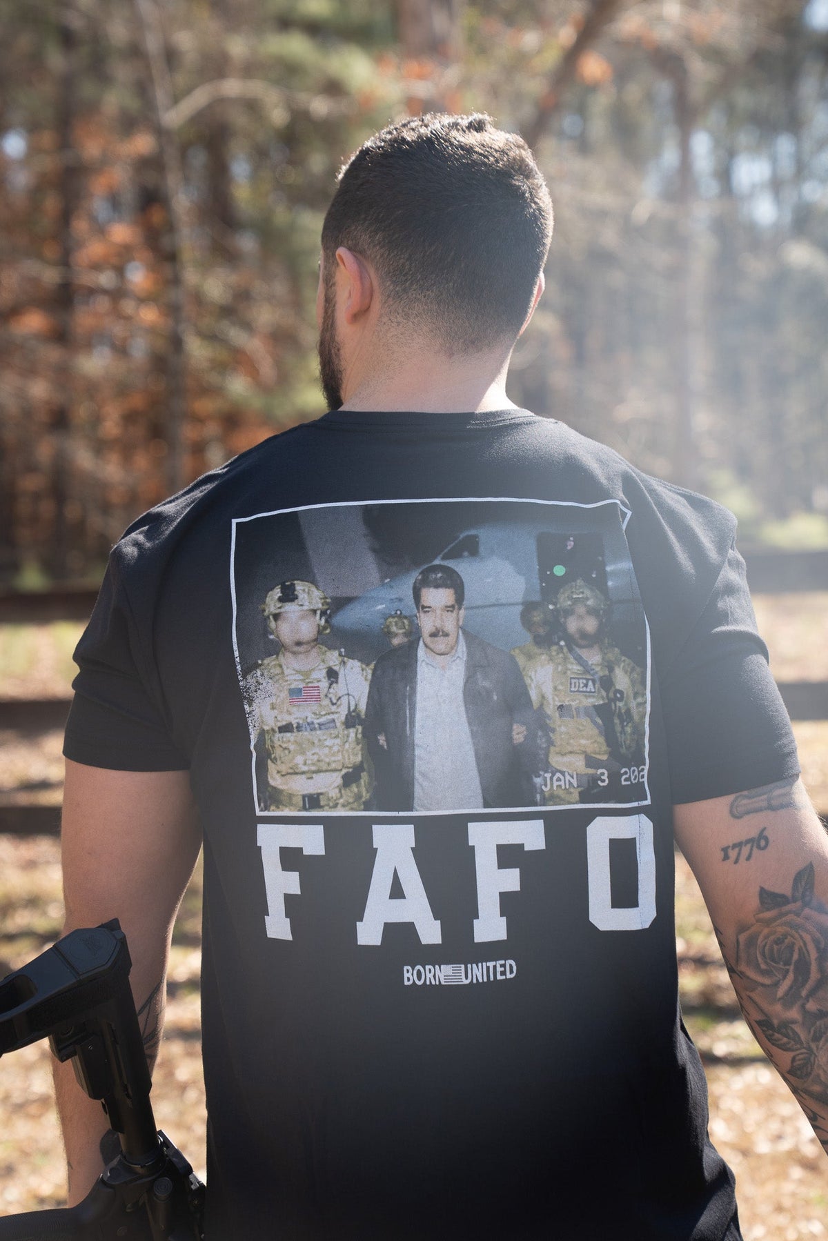 A man stands outdoors with his back to the camera, wearing the Born United Private Label FAFO - Maduro T-shirt, which features a photo of an arrest scene and bold FAFO and BORN UNITED text. Sunlight filters through the trees behind him.
