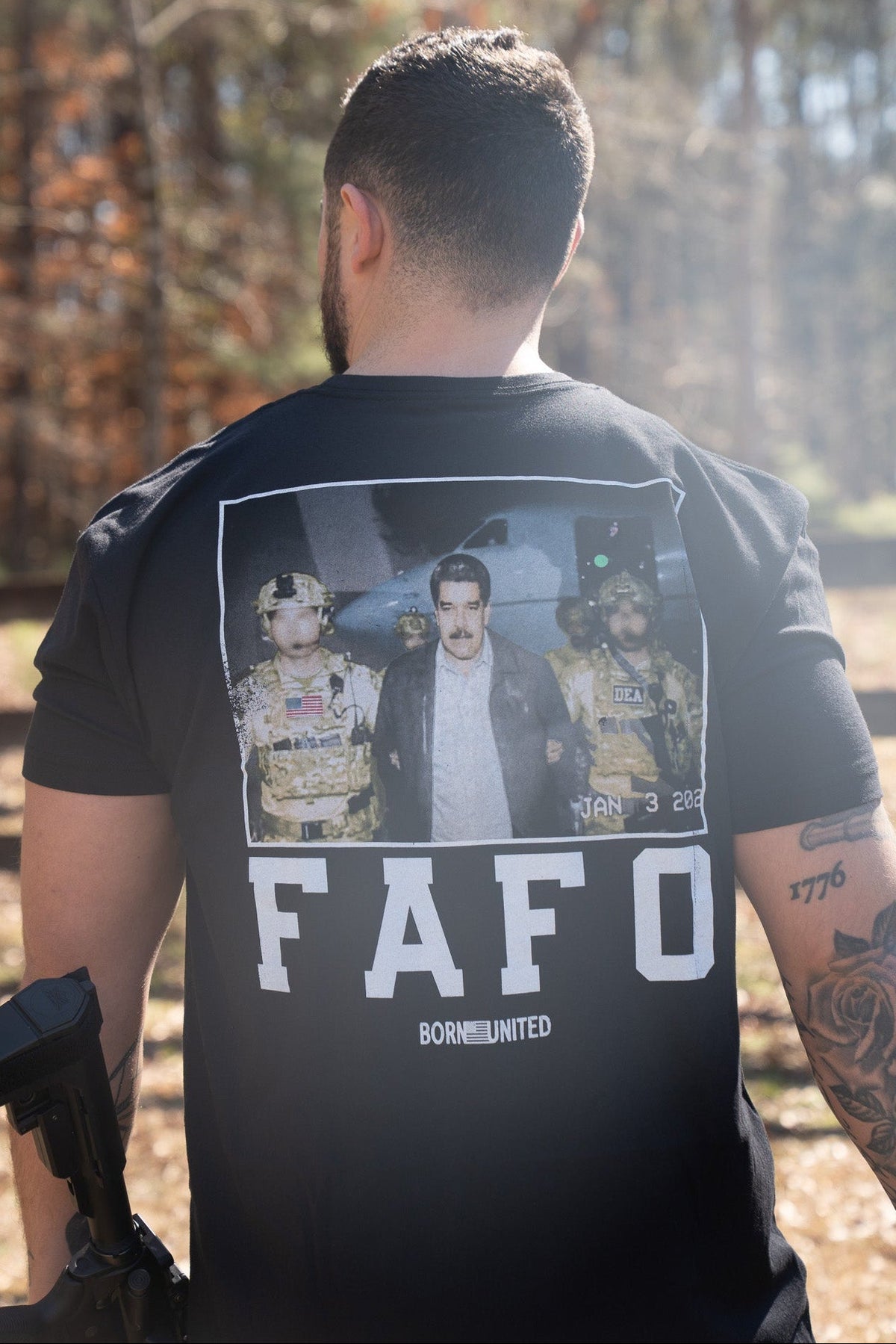 A man stands outdoors with his back to the camera, wearing the Born United Private Label FAFO - Maduro T-shirt, which features a photo of an arrest scene and bold FAFO and BORN UNITED text. Sunlight filters through the trees behind him.