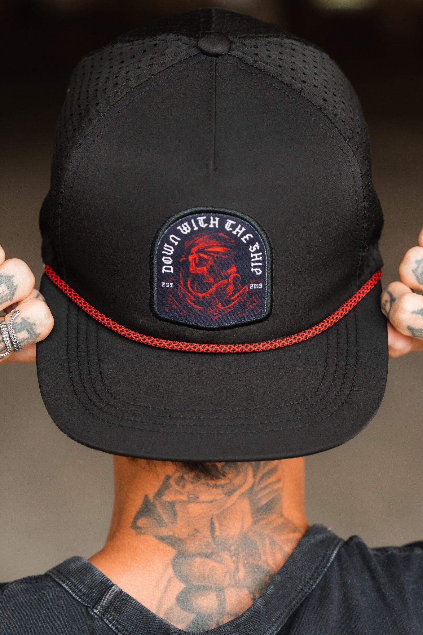 The image shows a person from behind wearing the Relentless Betrayal Ship Wreck Snapback - Red, a black cap with a red dragon patch and DOWN WITH THE SHIP in white. The brim features red rope detail, complementing their tattooed neck and hands for a unique style.