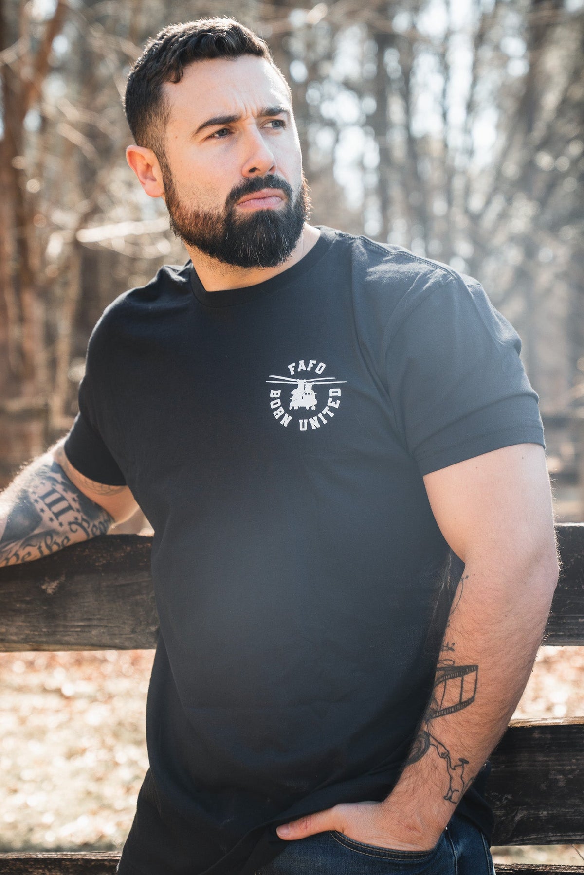 A bearded man with tattoos leans on a wooden fence outdoors, wearing a Born United Private Label FAFO - Maduro black t-shirt with a white logo; trees and sunlight brighten the background.