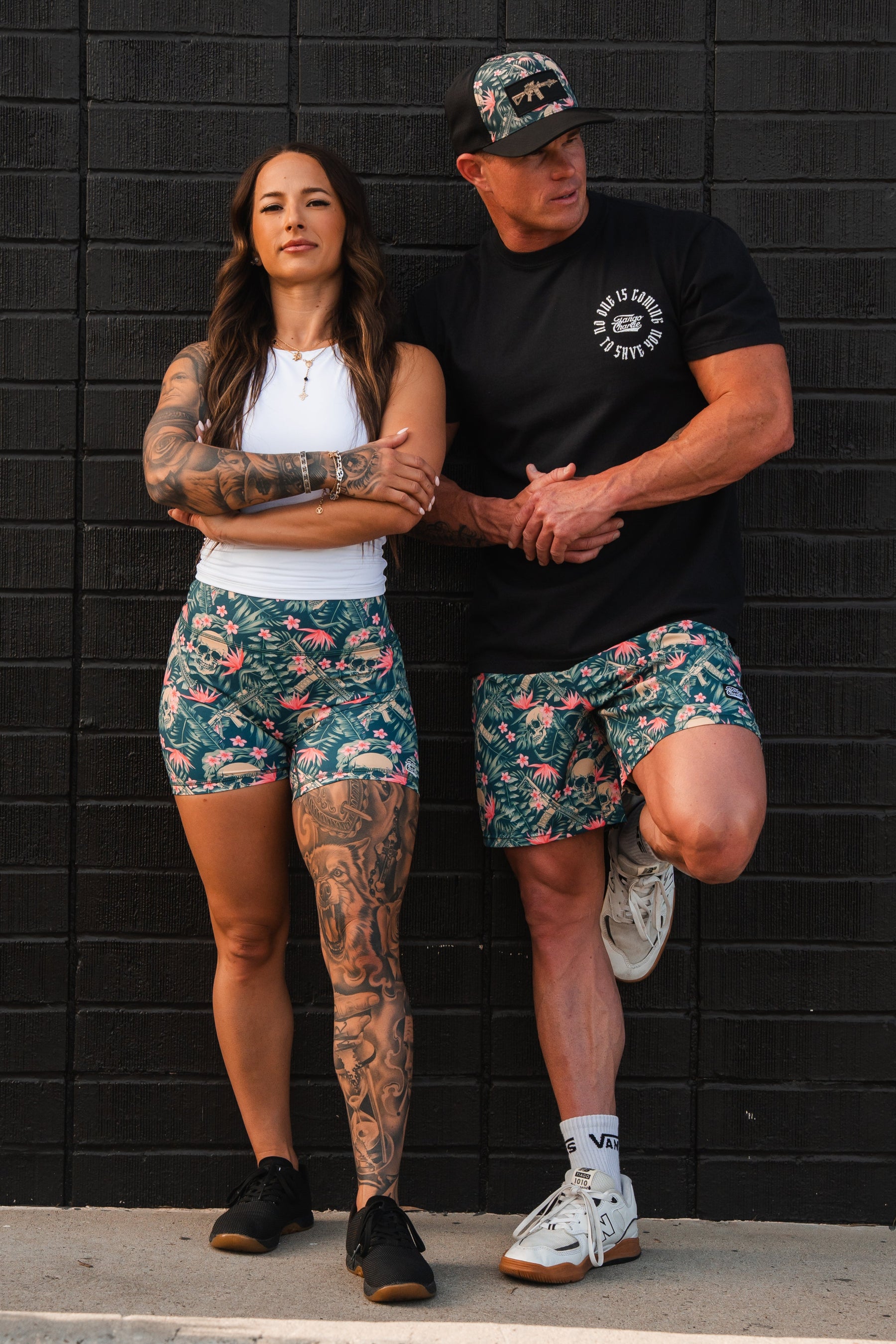 A woman and a man pose confidently against a black brick wall. Shes in Womens High-Waisted Seamless Front Compression Training Shorts by Tango Charlie Apparel, paired with a white tank top revealing her tattoos; hes wearing a black T-shirt and cap, both embodying an empowering design as they face the camera.