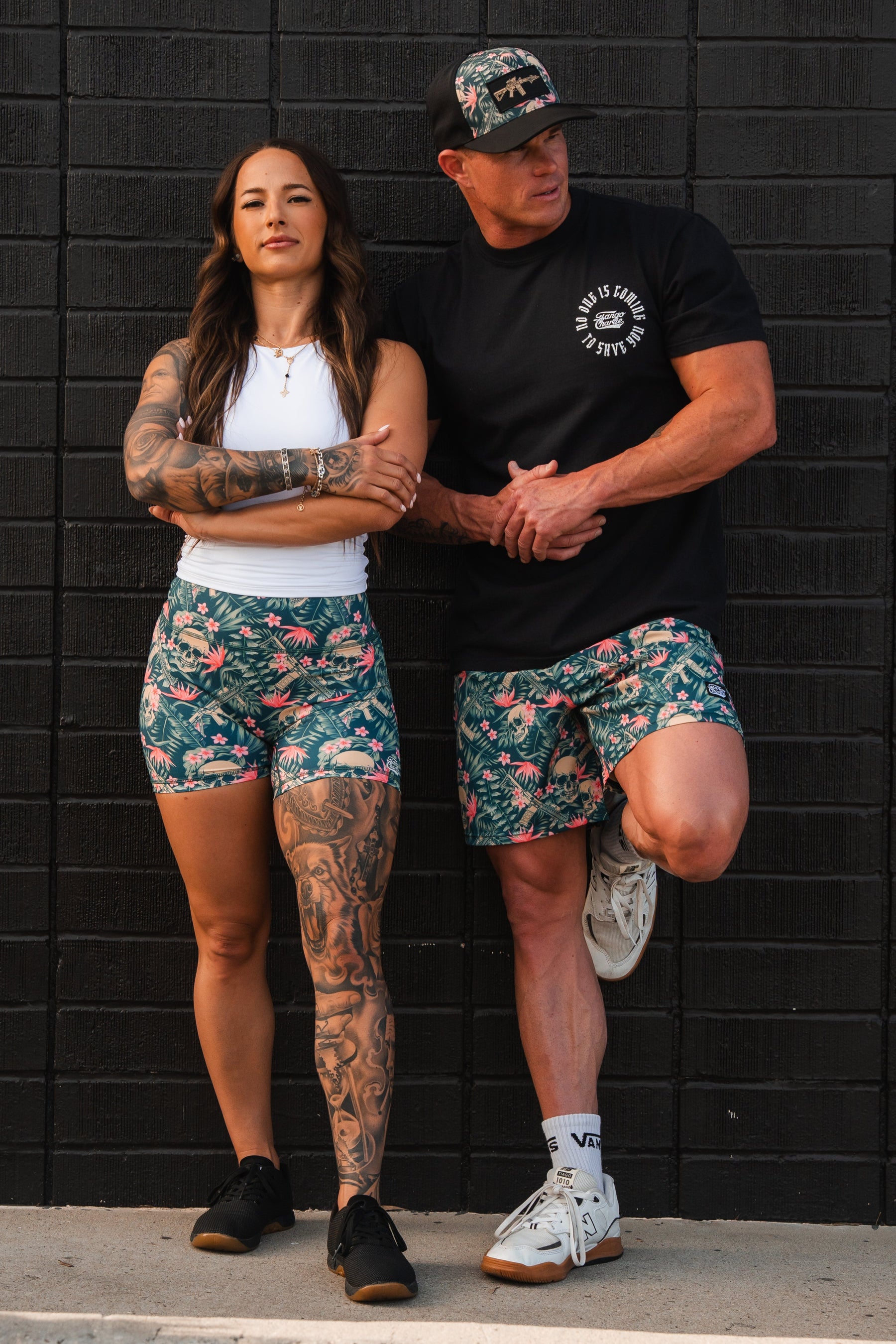 A woman in a white tank and floral shorts with a tattooed leg and a man in a black shirt, Tango Charlie Apparels Mens Performance Training Shorts 5.5 - Aloha AR, and a cap stand casually against a black brick wall, exuding confidence as they look ahead.