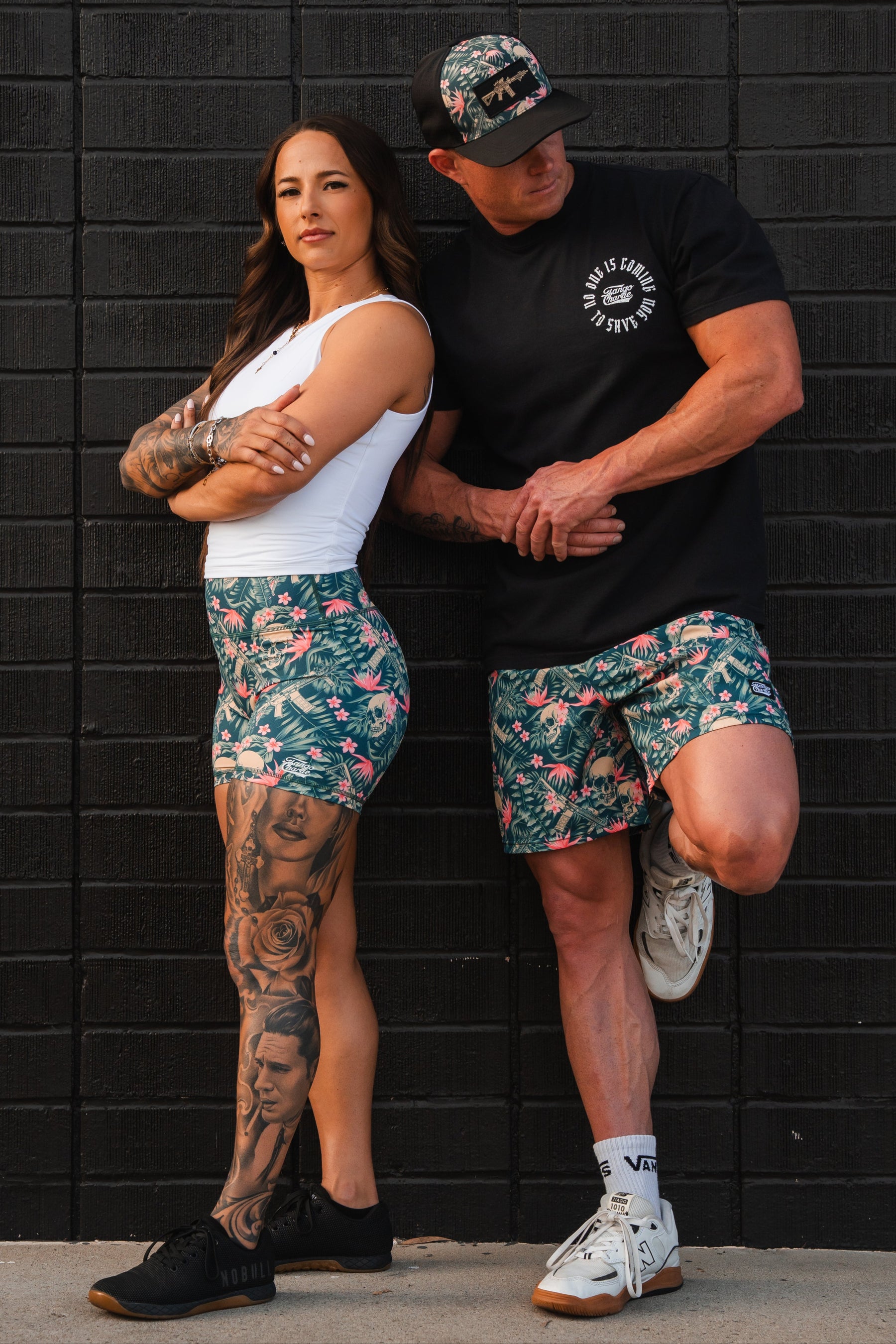 A woman and man pose against a black wall. She wears Tango Charlie Apparels empowering white tank, high-waisted seamless floral shorts, and black sneakers, showing tattooed legs. He exhibits an Aloha AR look with his black T-shirt, floral shorts, cap, and white sneakers. Both have athletic builds.