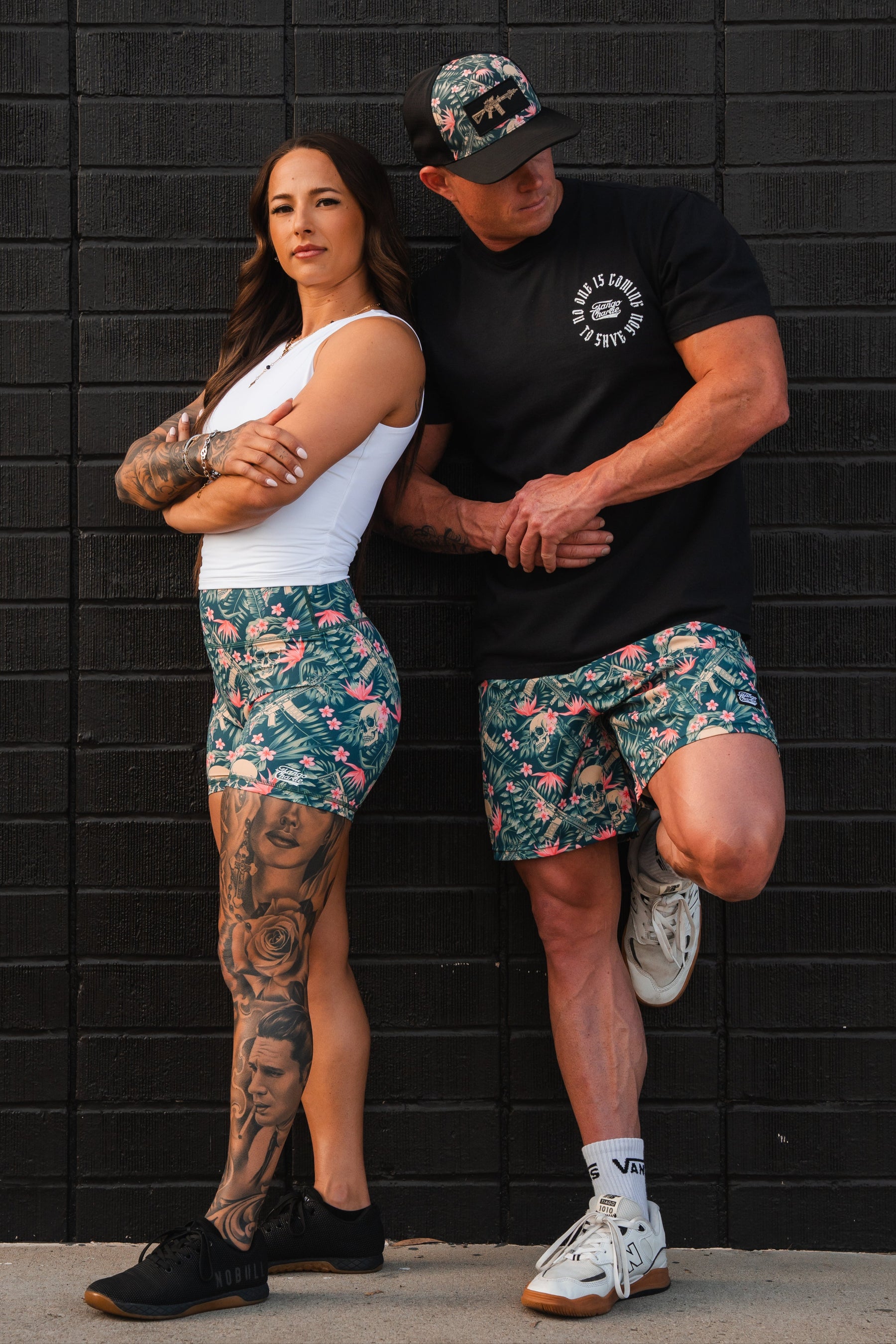 Against a black brick wall, a woman exudes confidence in her white top and floral Aloha AR Shorts, with tattoos visible on her leg. The man sports a Tango Charlie Apparel black shirt, Mens Performance Training Shorts 5.5 - Aloha AR, and a cap. Both strike relaxed poses, radiating casual coolness.