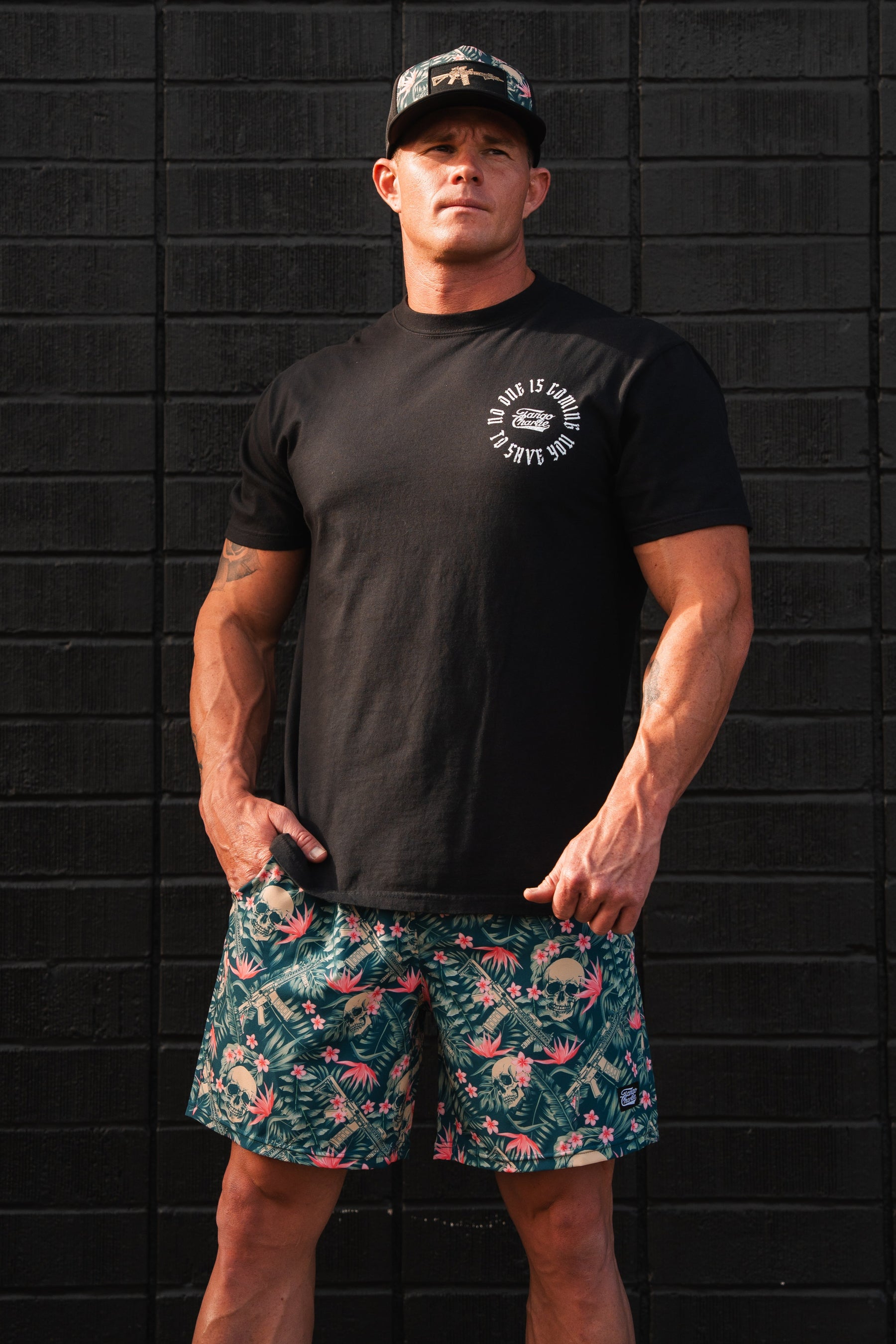 A man stands by a black brick wall wearing a black t-shirt and Tango Charlie Apparels Mens Performance Training Shorts | 5.5- Aloha AR with floral patterns. Hes tattooed and capped, with his hands relaxed by his sides.