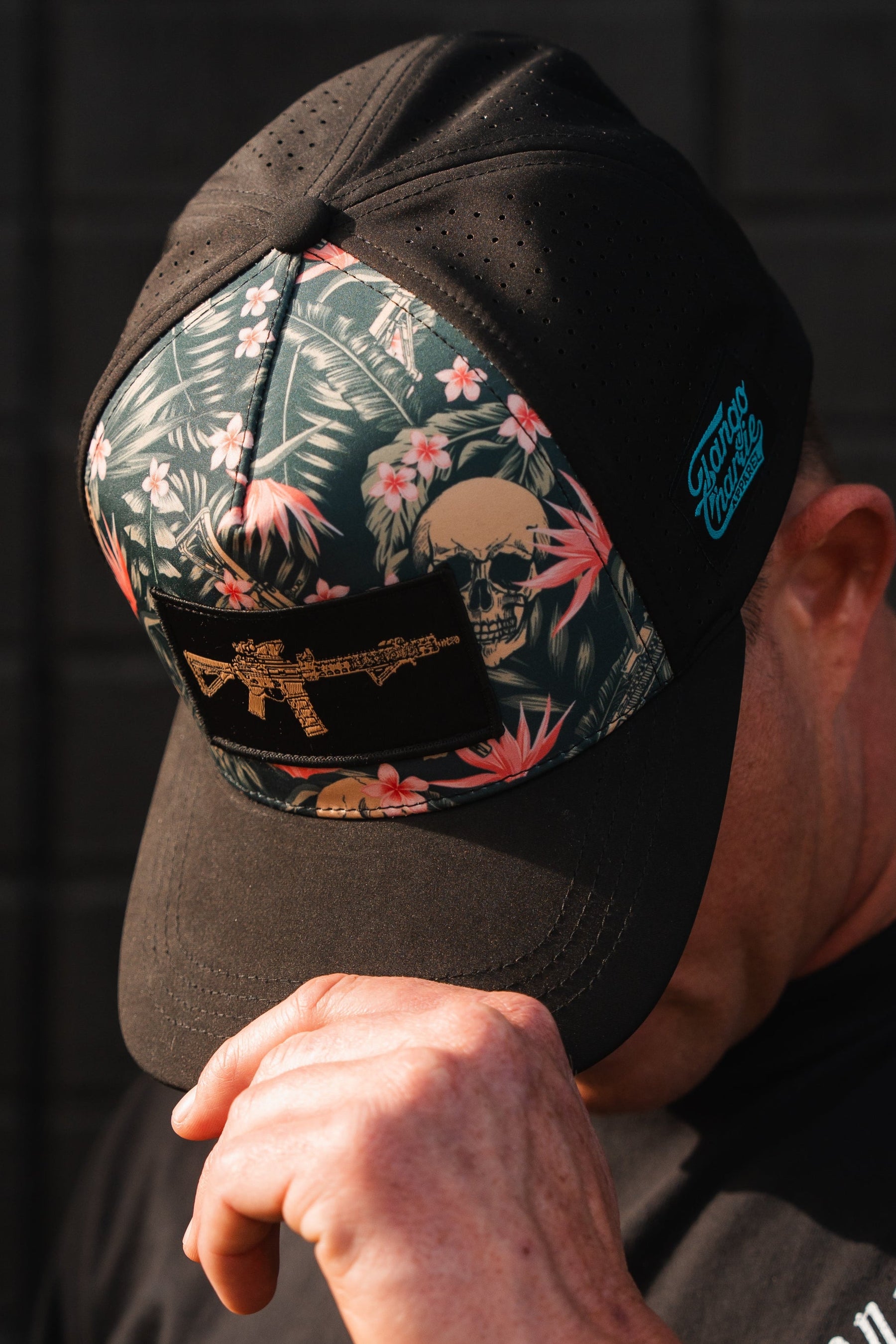 A man wears the Tango Charlie Apparel Aloha AR Hydro snapback hat, featuring a black cap with a floral and skull design, a gold gun graphic, and a blue logo on the side. The water-resistant hat offers breathability for comfort as he tilts his head forward, touching its brim with his right hand.