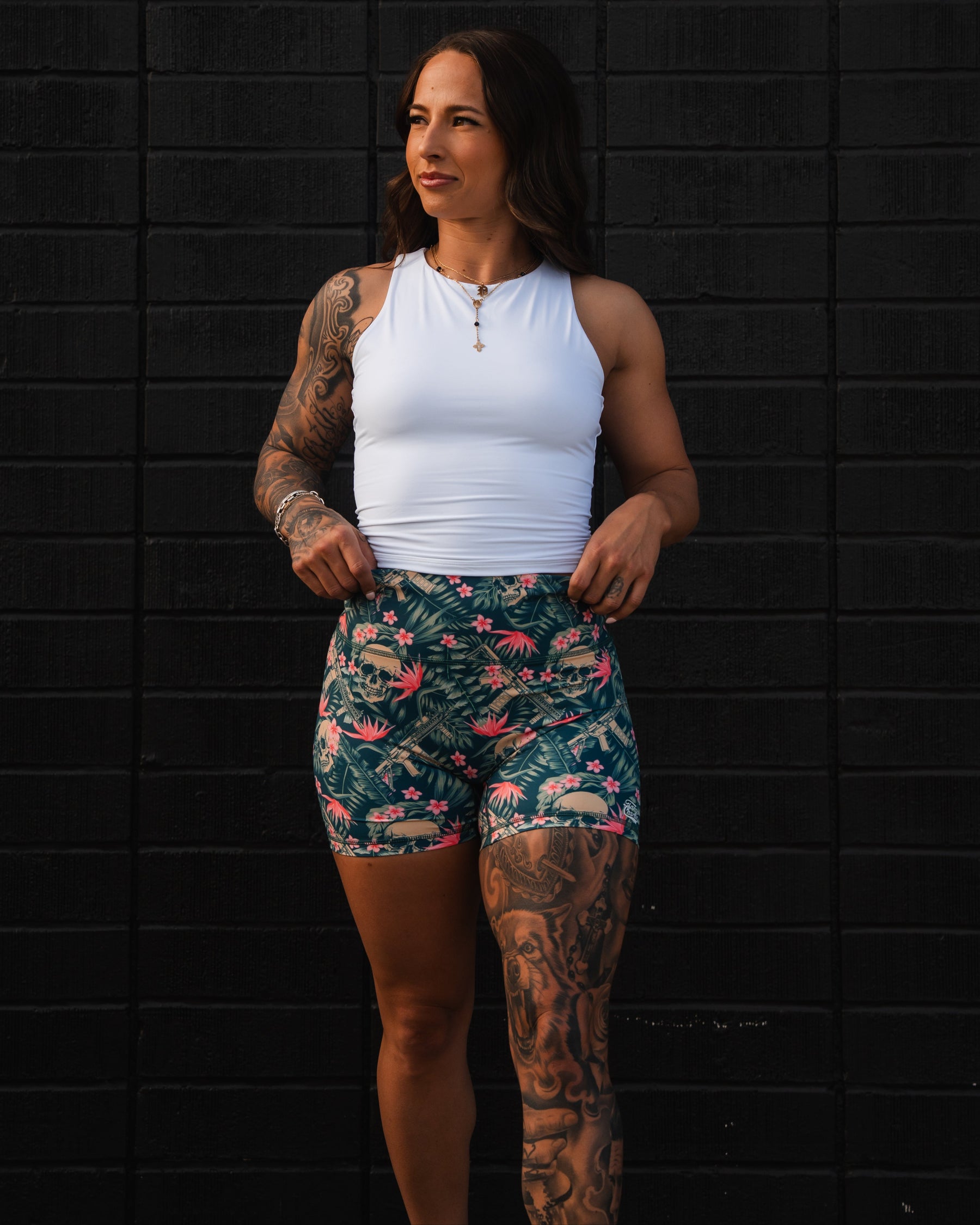 A woman in a white tank top and Tango Charlie Apparels Womens High-Waisted Seamless Front Compression Training Shorts | Aloha AR poses confidently against a black brick wall. Her flowing hair and tattoos showcase an empowering design amid dramatic lighting.