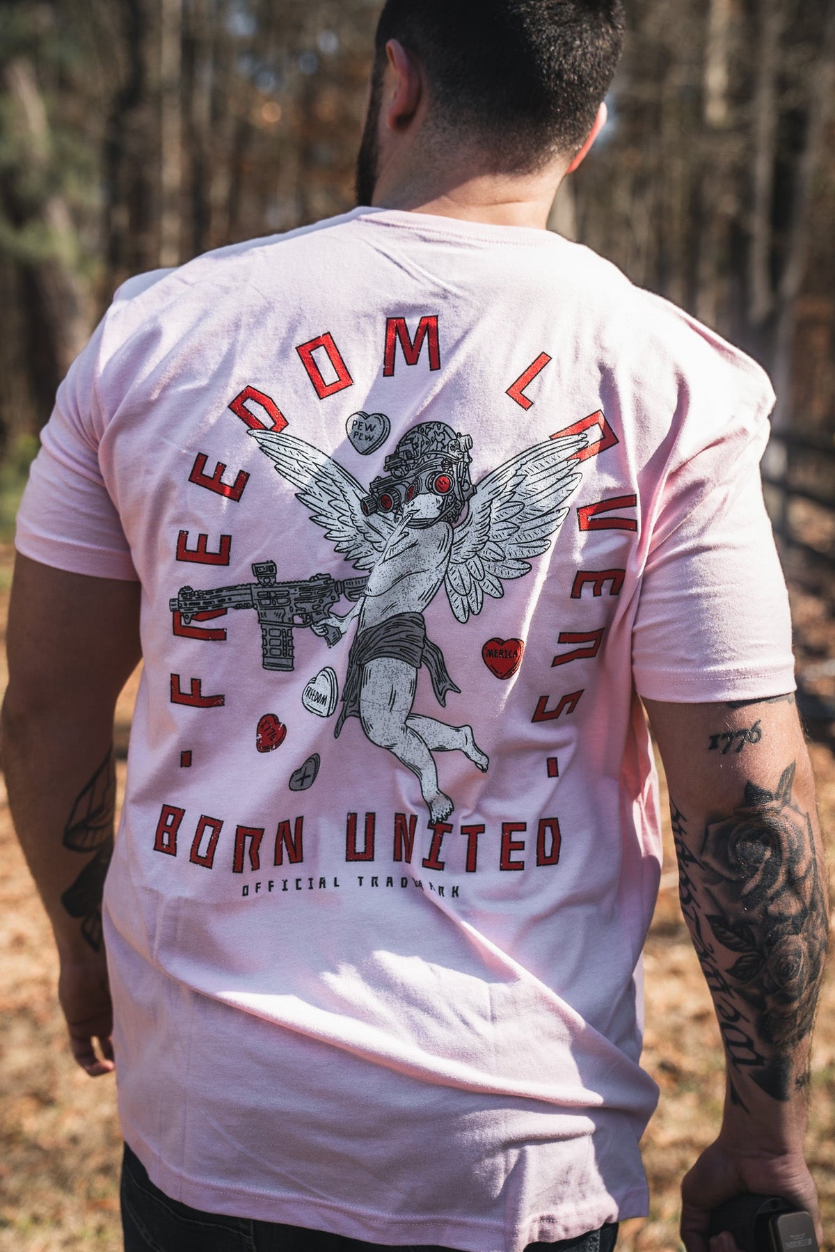 A man with tattooed arms wears the Cupid’s Shot V2 tee by Born United Private Label—a light pink shirt with a tactical cherub and rifle graphic, plus “Freedom Layer” and “Born United” on the back. Trees and fencing are behind him.