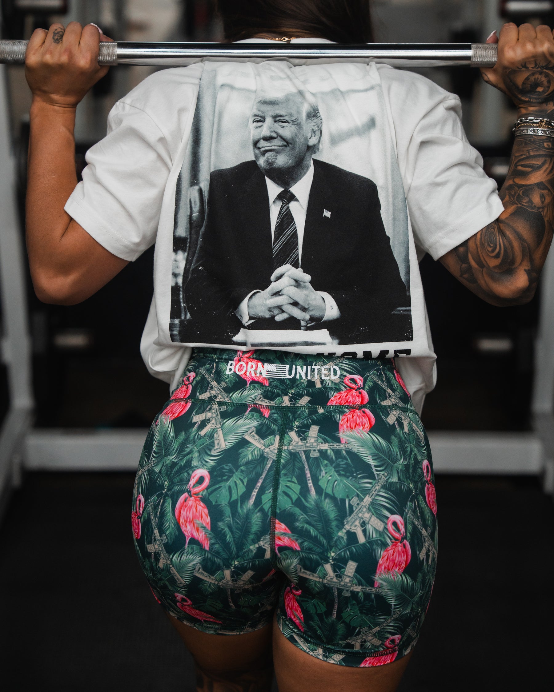 Seen from behind in a gym, a person lifts weights wearing a T-shirt featuring a black-and-white image of a man in a suit and Womens High-Waisted Seamless Front Compression Training Shorts by Born United Private Label. These shorts are adorned with flamingos and rifles, offering stretchable comfort with every lift.