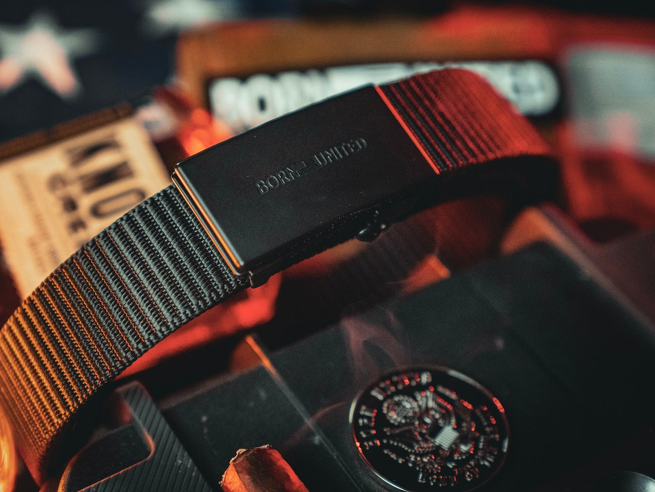 A close-up of the BU Logo Belt - Black from Born United Private Label, featuring the engraved logo on the buckle, with blurred patriotic-themed objects and warm lighting in the background.