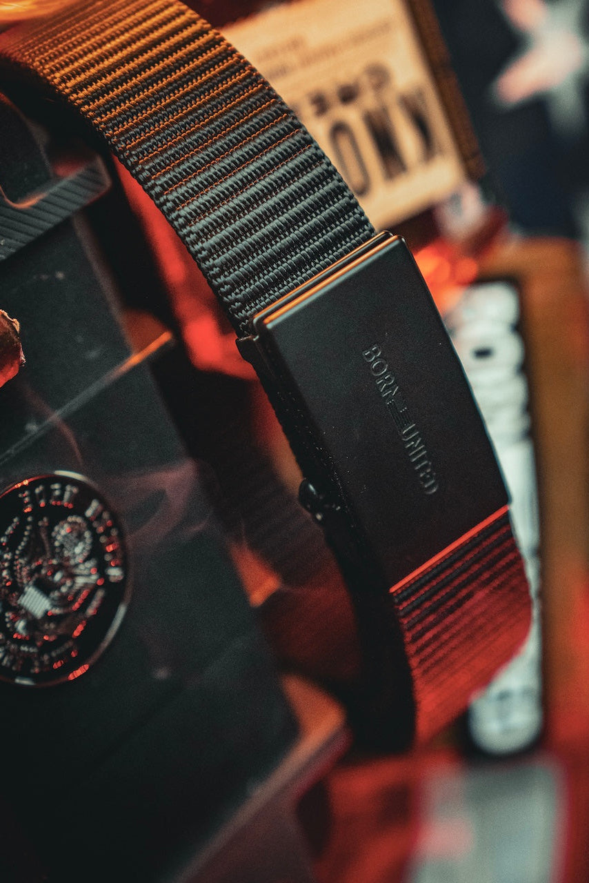 A close-up of the BU Logo Belt - Black from Born United Private Label, featuring the engraved logo on the buckle, with blurred patriotic-themed objects and warm lighting in the background.