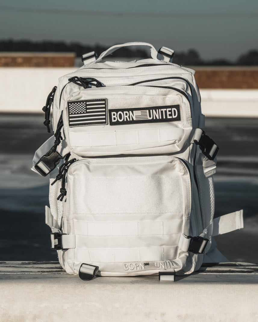 The Storm Trooper White Premium Tac-Pac - 25L by Born United Private Label, featuring multiple compartments and an American flag patch with BORN UNITED, rests on a concrete ledge against a clear blue sky, standing out amidst an urban rooftop scene.