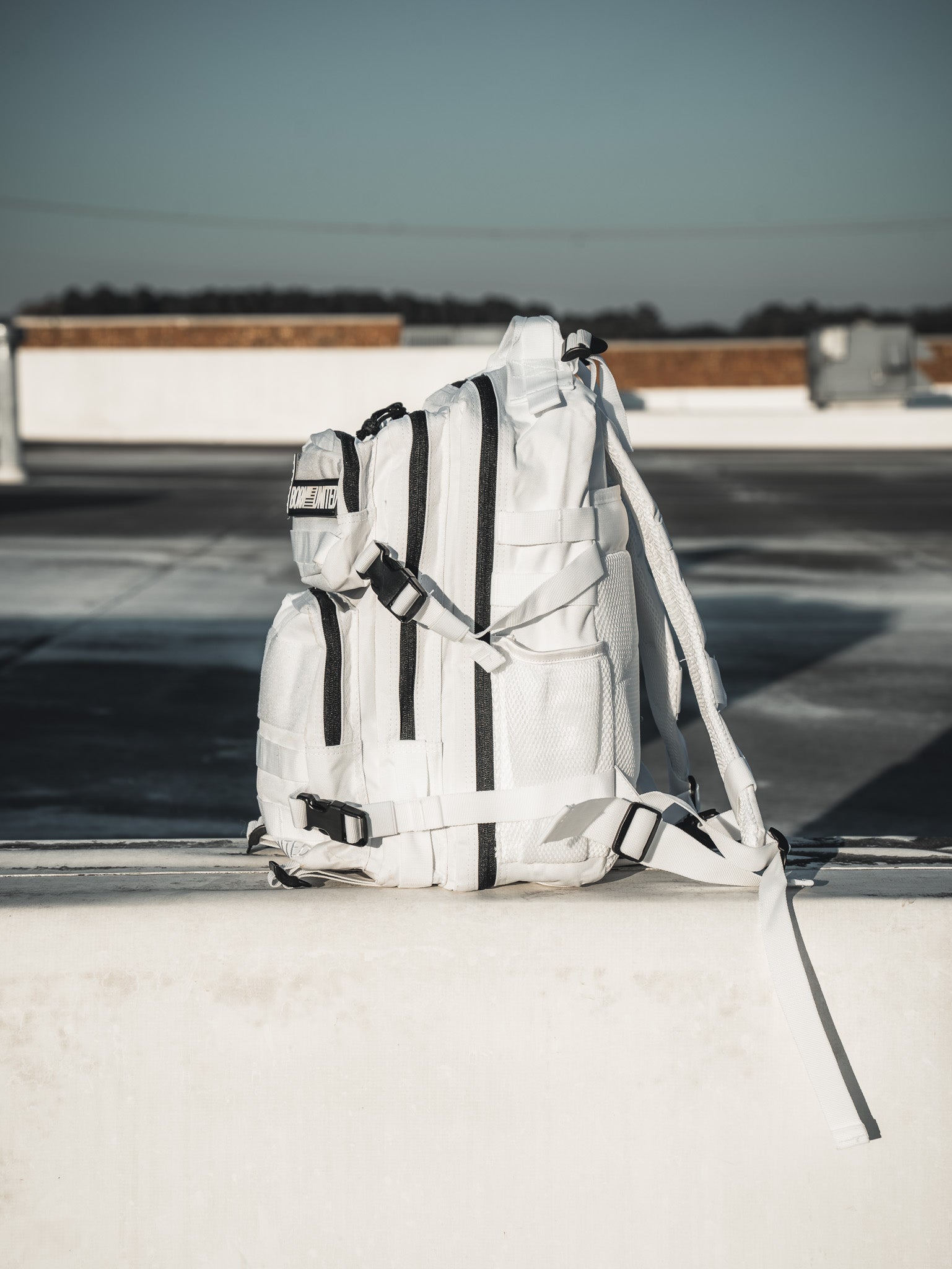 The Born United Private Labels Storm Trooper White Premium Tac-Pac - 25L, featuring black straps, is set on concrete with a rooftop backdrop. Bright lighting casts shadows, highlighting its details.