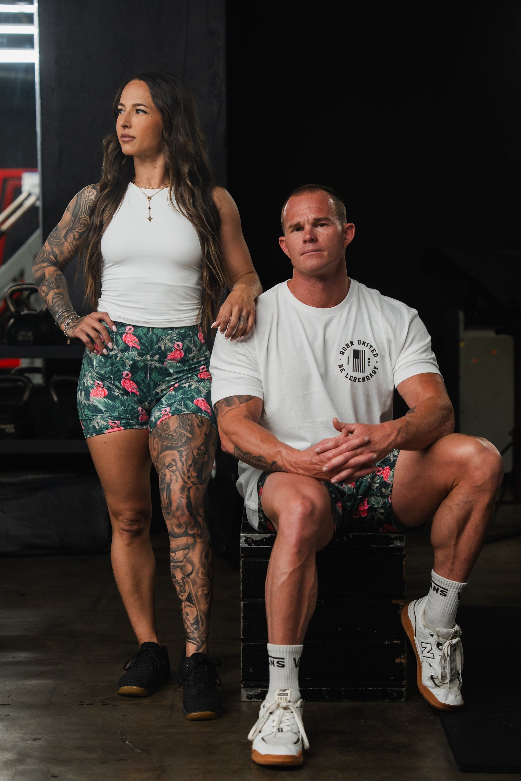 In the gym, a woman in Born United Private Labels Womens High-Waisted Seamless Front Compression Training Shorts in Loaded Flamingo stands beside a man seated on a box. She rests her hand on his shoulder while he wears a white t-shirt and breathable shorts, both exhibiting athletic builds with tattoos.