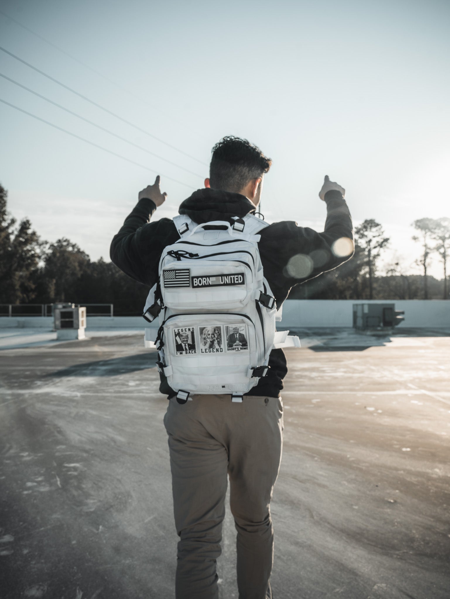 A person with short dark hair stands on a rooftop, sporting the Born United Private Labels Storm Trooper White Premium Tac-Pac - 25L. The sun is low, casting a lens flare as they give double thumbs up.