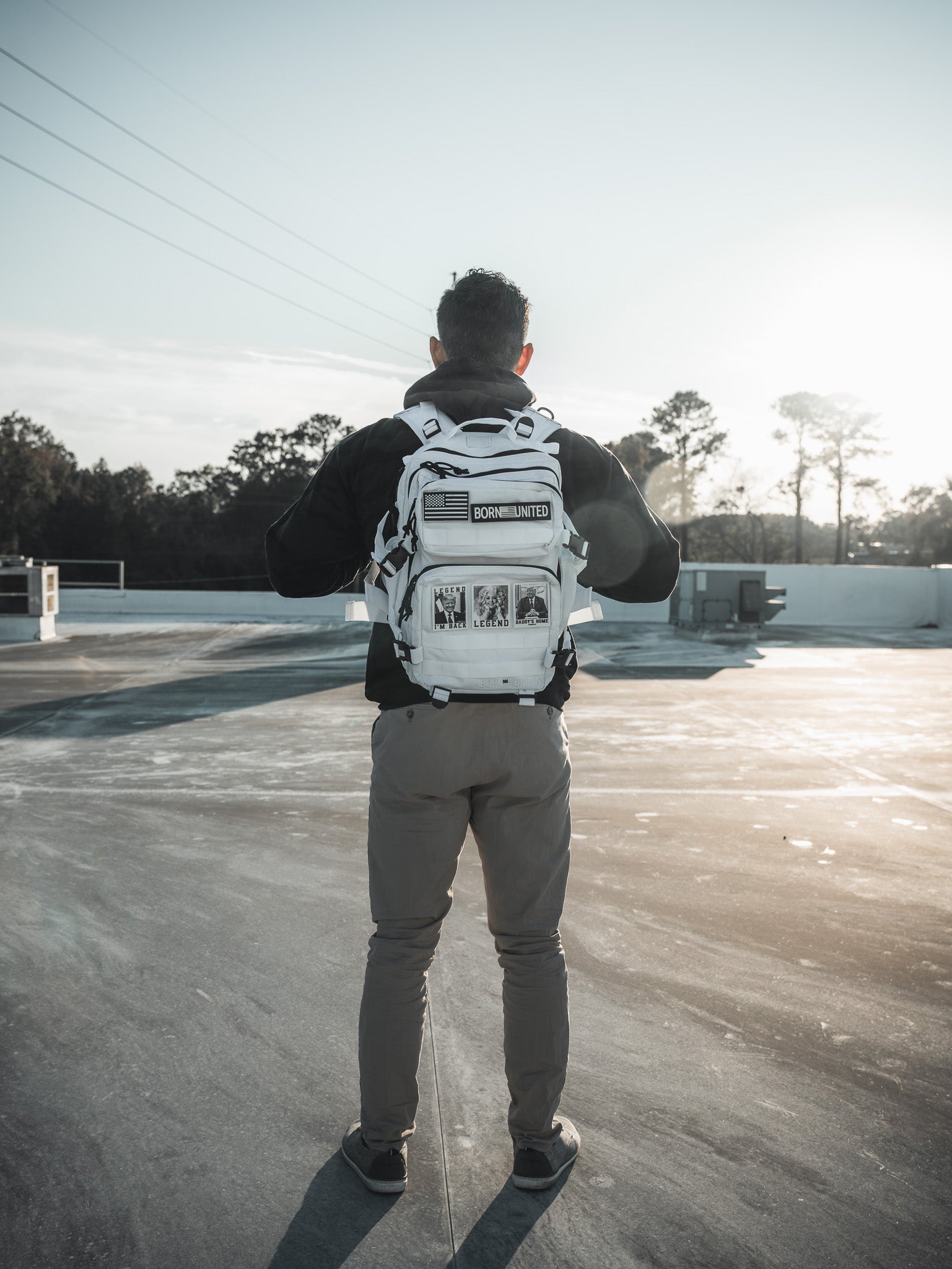 On a sunlit rooftop, someone in a black jacket and gray pants stands with their back turned, showcasing the Born United Private Label Storm Trooper White Premium Tac-Pac - 25L. Trees and a clear sky create the perfect backdrop for this standout accessory.