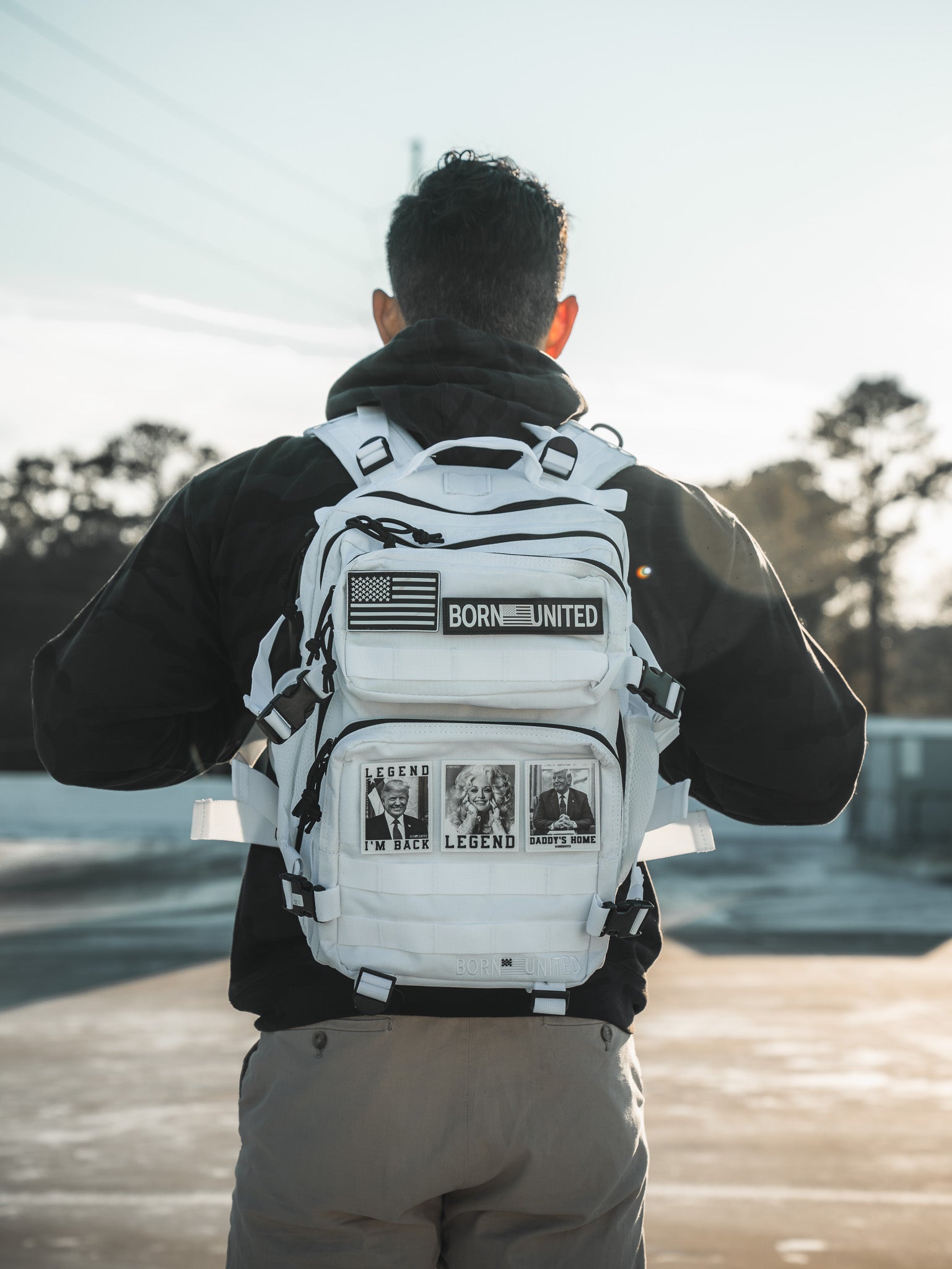 Standing outdoors in a dark jacket and khaki pants, the person faces away with the Storm Trooper White Premium Tac-Pac - 25L by Born United Private Label. Adorned with patches like an American flag and Legend photos, the backpack steals attention against a tree-lined sky.