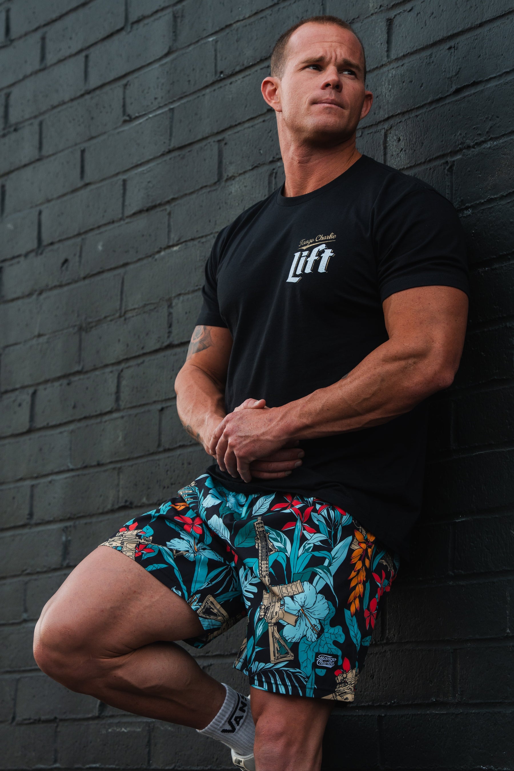 A man in a black T-shirt and colorful shorts leans against a dark brick wall, arms crossed, gazing sideways. His Mens Performance Training Shorts | 5.5- Tactical Tropic by Tango Charlie Apparel feature tropical leaves and parrots for a stylish yet versatile design.