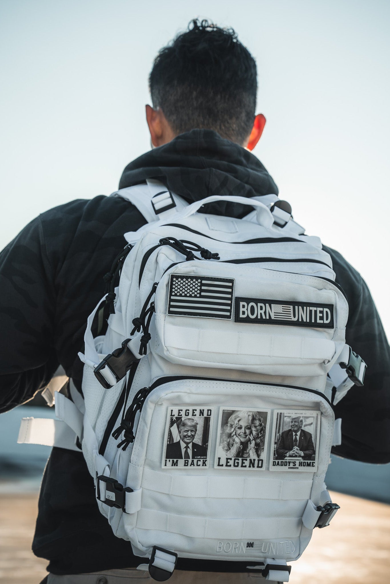 A person in a black hoodie faces away, carrying the Born United Private Label Storm Trooper Tac-Pac - 25L backpack, decorated with patches such as the U.S. flag, BORN UNITED, and black-and-white LEGEND, IM BACK, and DADDYS HOME images.