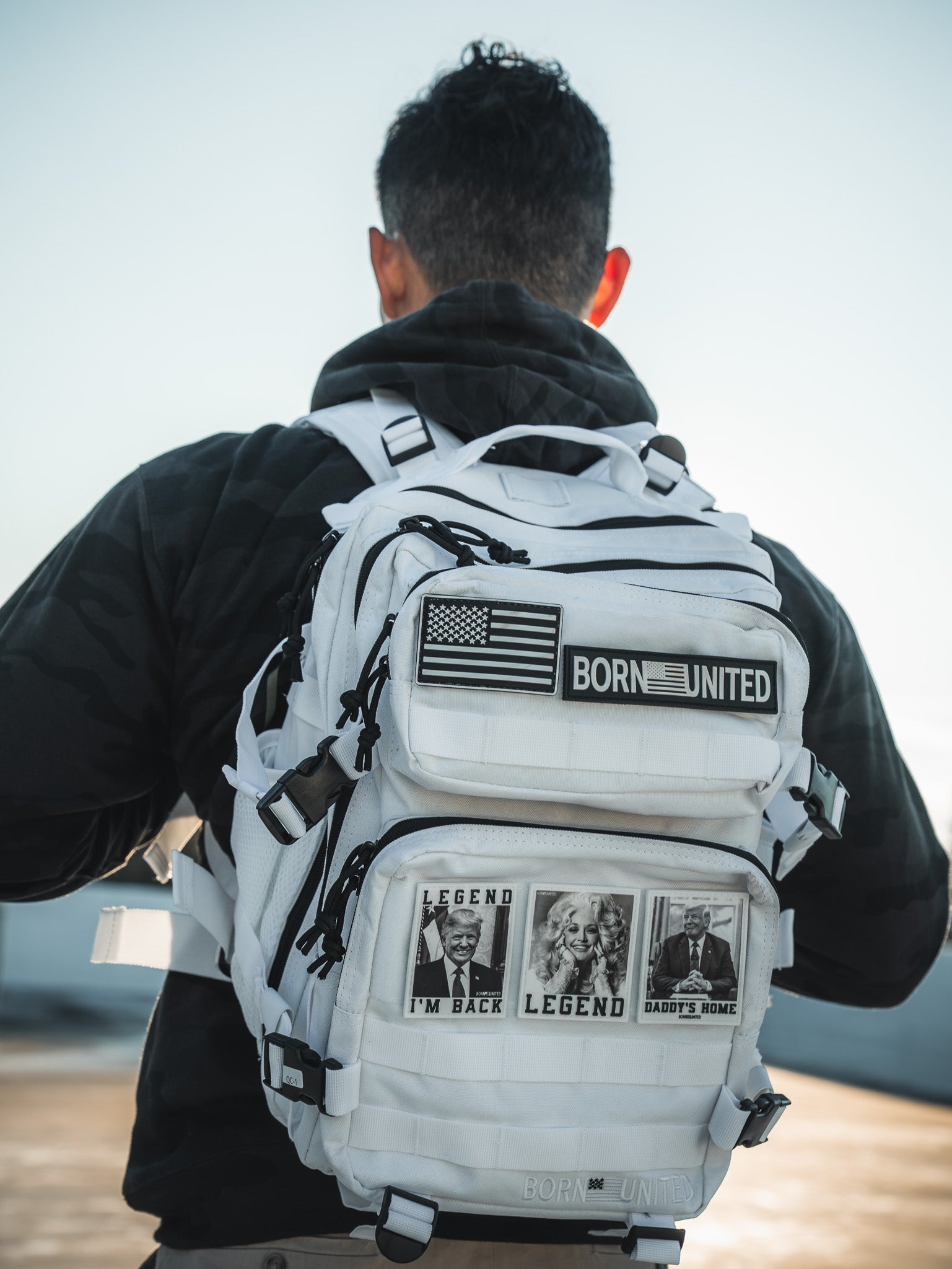 A person with short hair in a dark jacket carries Born Uniteds Storm Trooper White Premium Tac-Pac - 25L, adorned with patches like an American flag and photos, resembling a lone storm trooper as they stand outdoors against a light sky.