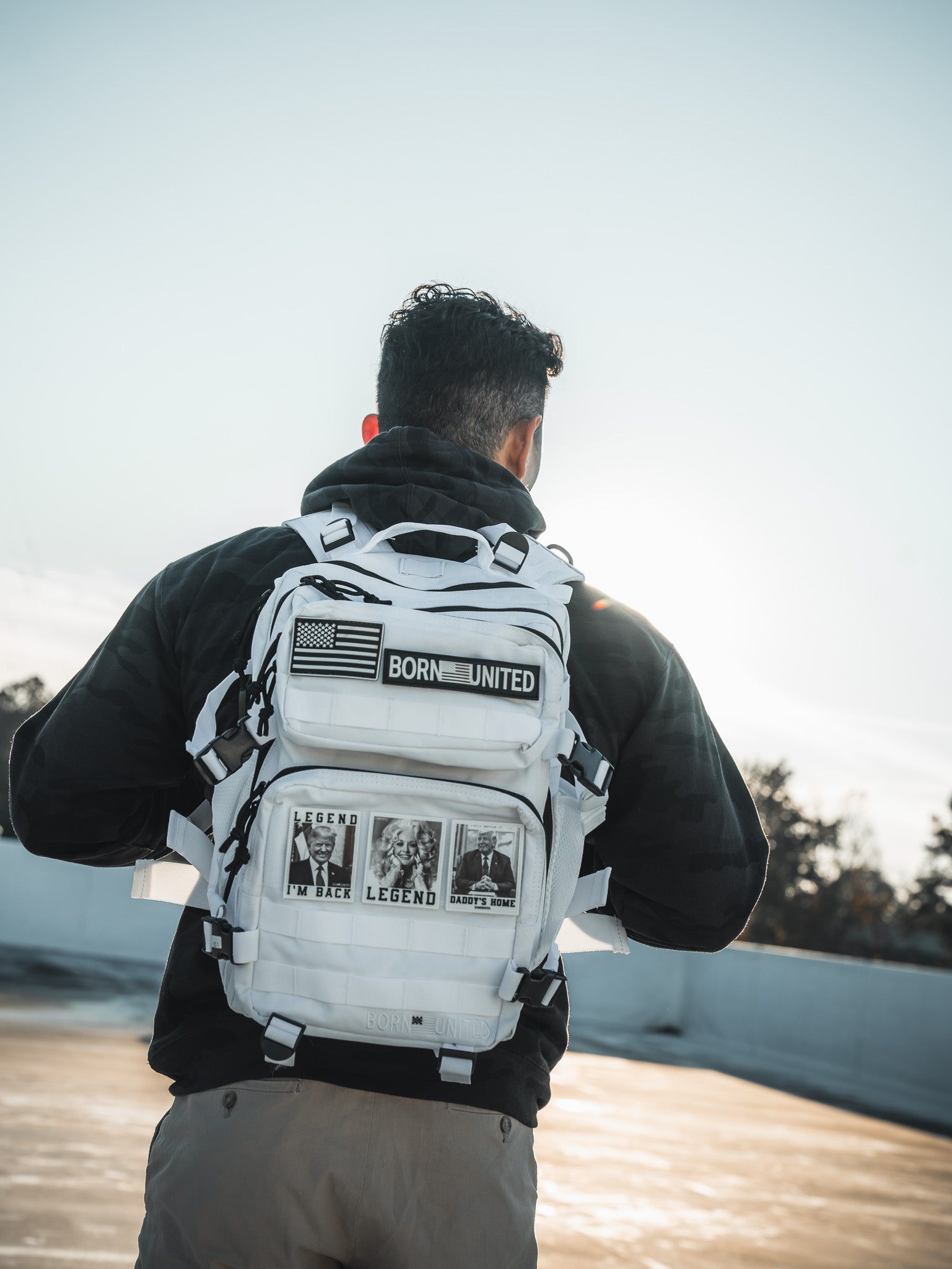A person with short dark hair stands outside in a black jacket and beige pants, wearing a Storm Trooper White Premium Tac-Pac 25L by Born United Private Label. The white backpack is adorned with patches, including an American flag and BORN UNITED. They face away, framed by clear skies and vibrant trees.