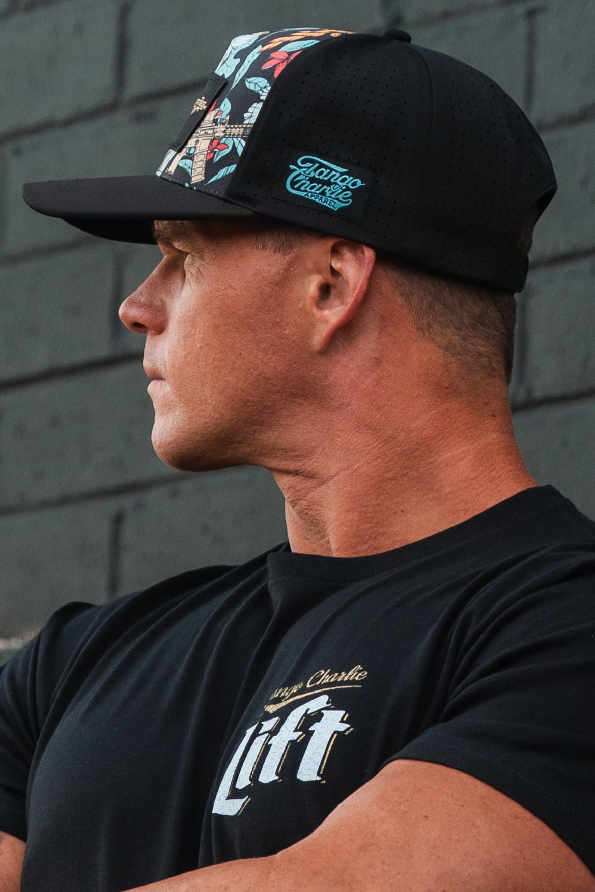 A person wearing a Tactical Tropics Hydro - Snapback Hat by Tango Charlie Apparel and a Tactical Tropics T-shirt stands in profile against a gray brick wall, looking to the side.