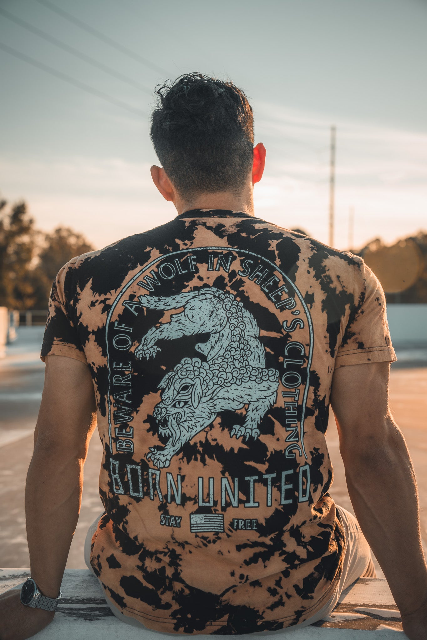 As the sun sets, a person sits on a rooftop wearing the Beware - Bleach Wash tie-dye shirt. Featuring a wolf and the words Beware of savage wolves in sheeps clothing, its from Born United Private Label. Trees and a clear sky stretch into the horizon.