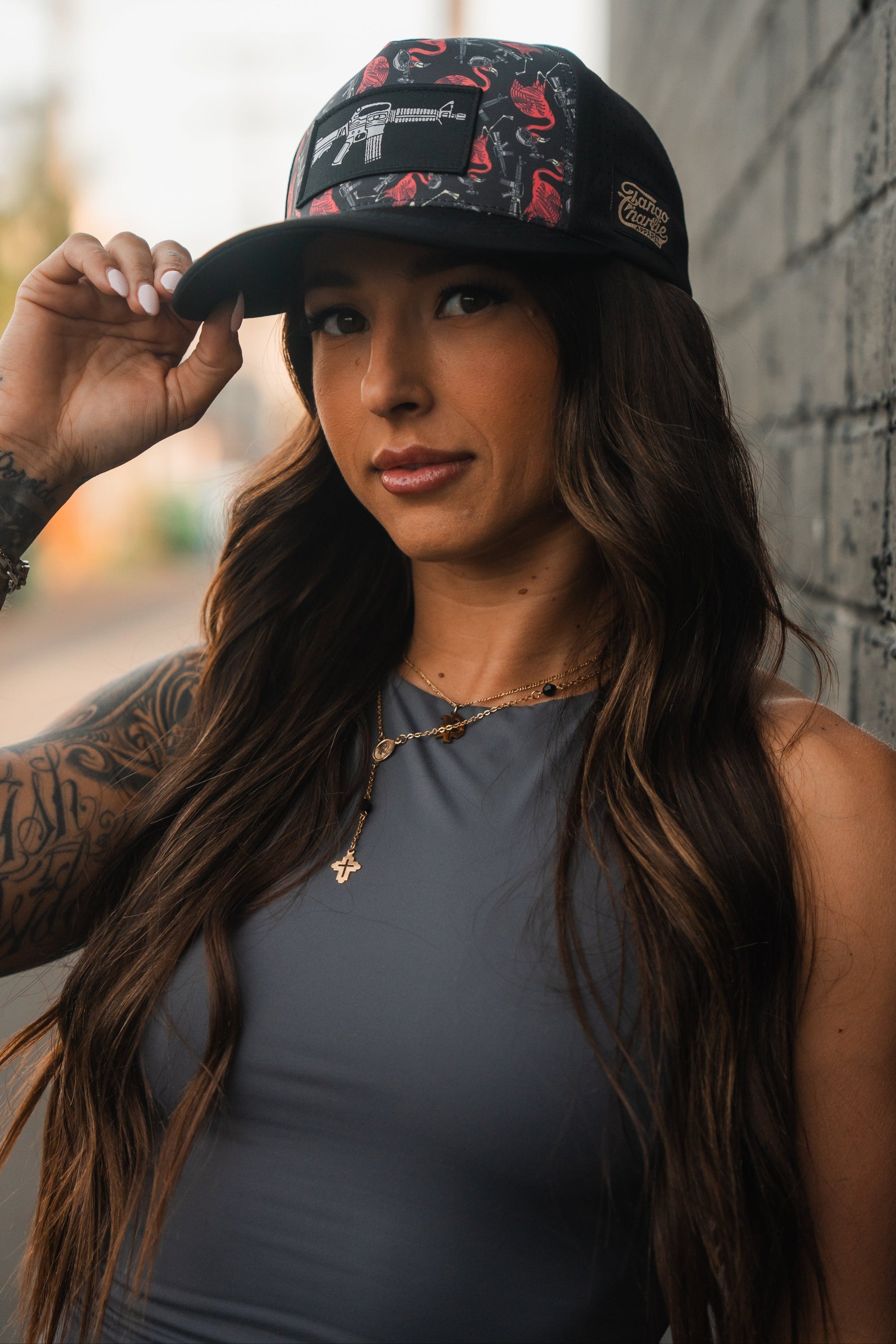 A woman wearing the Tactimingo Hydro Snapback Hat by Tango Charlie Apparel, featuring a subtle red pattern, and a sleeveless gray top, stands against a brick wall. With long brown hair and tattoos on her left arm, she accessorizes with a cross necklace while adjusting her cap confidently.