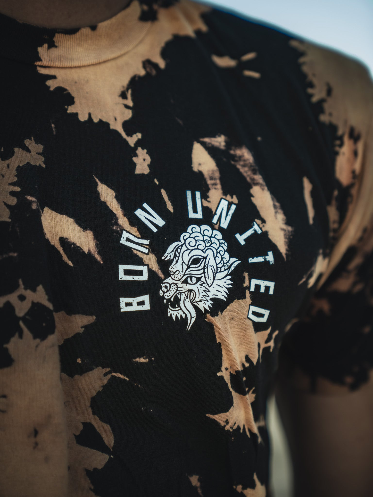 Close-up of someone in a black and tan tie-dye Beware - Bleach Wash t-shirt from Born United Private Label, featuring Born United above a stylized wolf graphic.