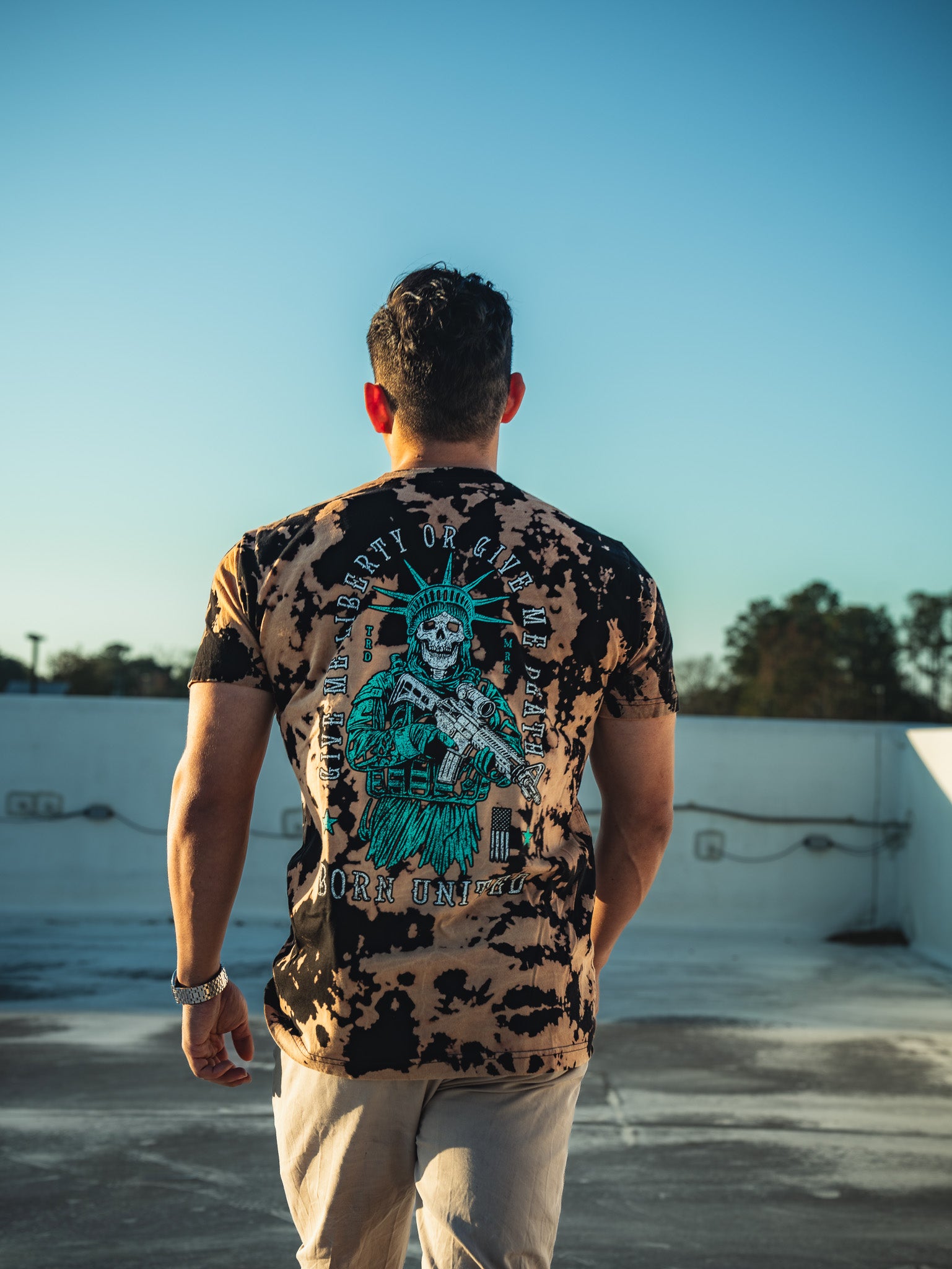 On a rooftop against a clear sky, a man dons Born Uniteds Liberty or Death - Bleach Wash T-shirt, featuring a skeleton Statue of Liberty with a gun and the words LIBERTY OR DEATH and FOR UNITY, capturing the essence of freedom for modern patriots.