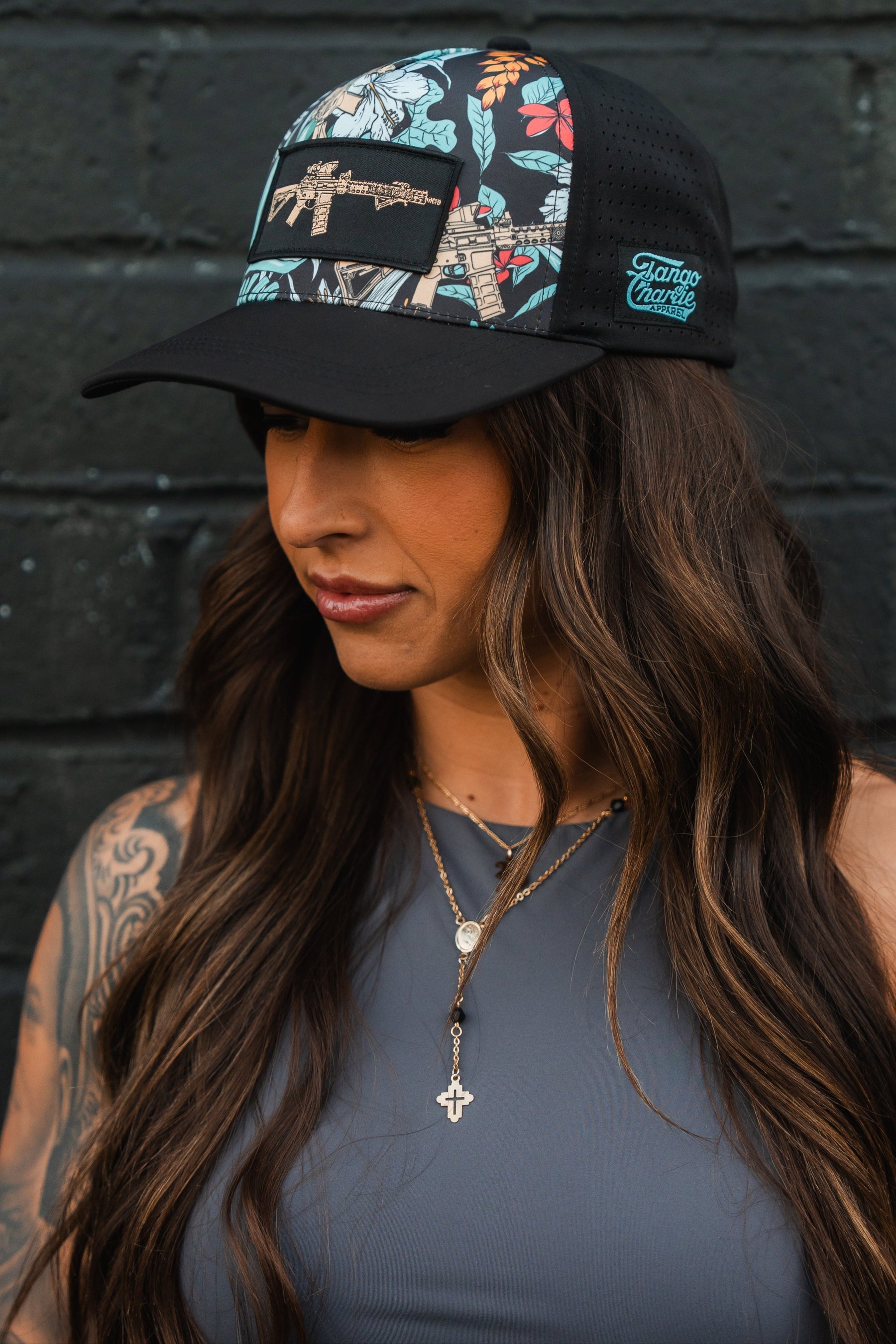 A woman with long brown hair is seen in a Tactical Tropics Hydro - Snapback Hat by Tango Charlie Apparel, boasting colorful patterns and a snapback closure. Dressed in a gray sleeveless top and layered necklaces with pendants, she stands against a black brick wall, gazing down to her right.
