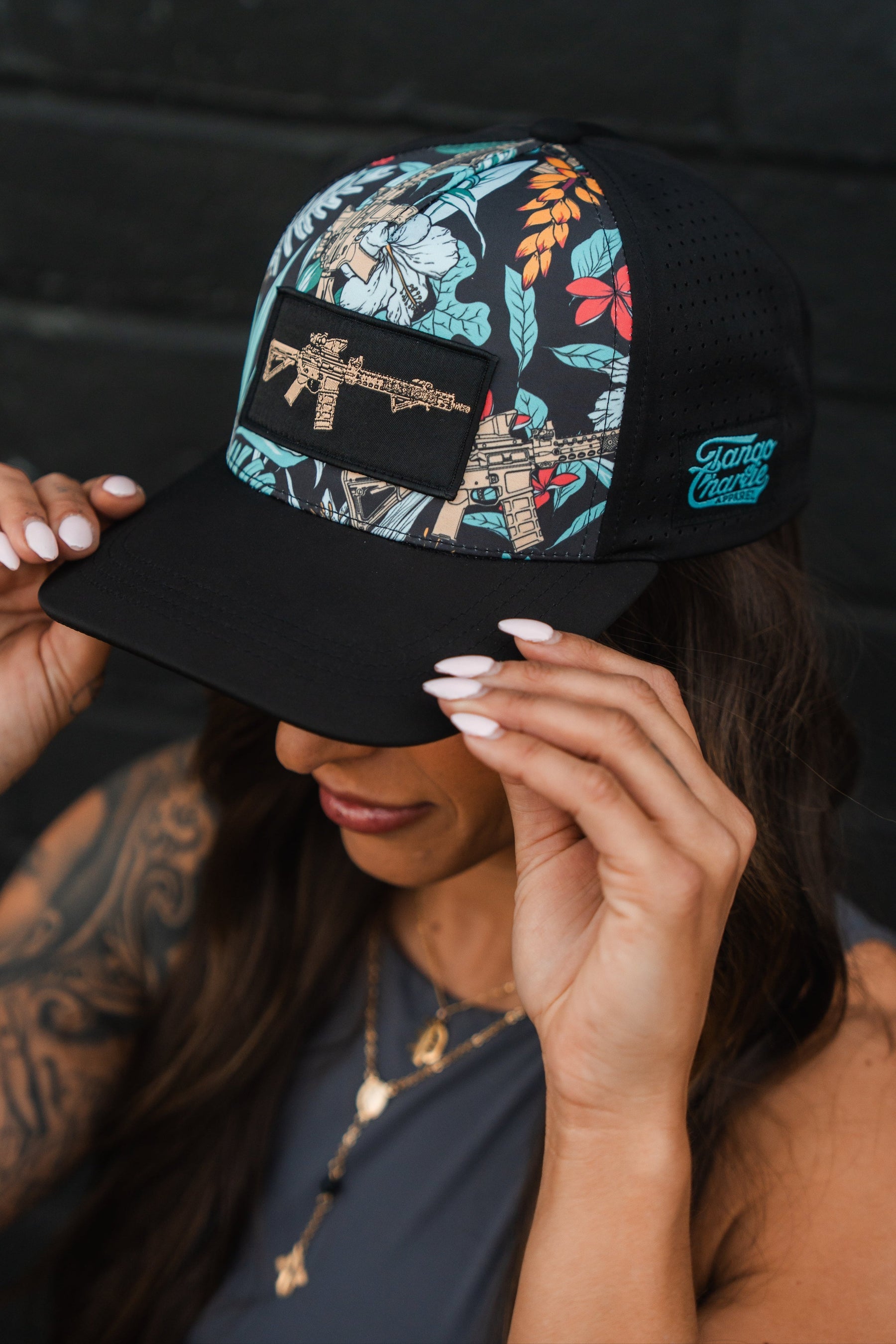 A woman with long hair and tattoos adjusts her Tango Charlie Apparels Tactical Tropics Hydro Snapback Hat, featuring a black floral design with an embroidered gun. With manicured white nails and layered necklaces, she stands against a dark wall. The hat is hydro-resistant and offers comfort with its snapback enclosure.